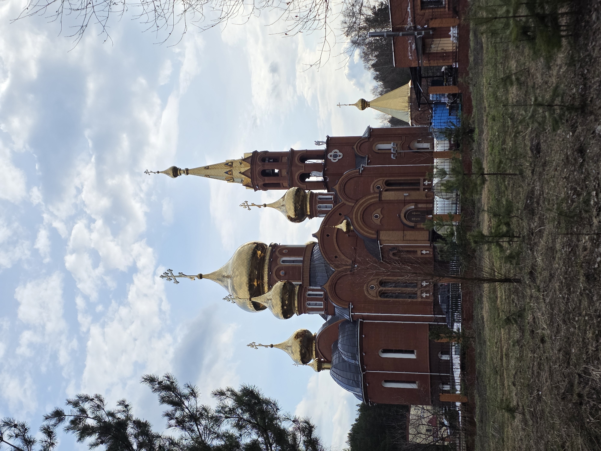 The image shows the Church of St. George the Victorious, located in Chaikovsky, Russia. The church is constructed of red brick and features multiple golden domes with crosses. The surrounding environment includes trees, grass, and a fence. The sky is visible in the background, with scattered clouds. There are no people visible in the image.