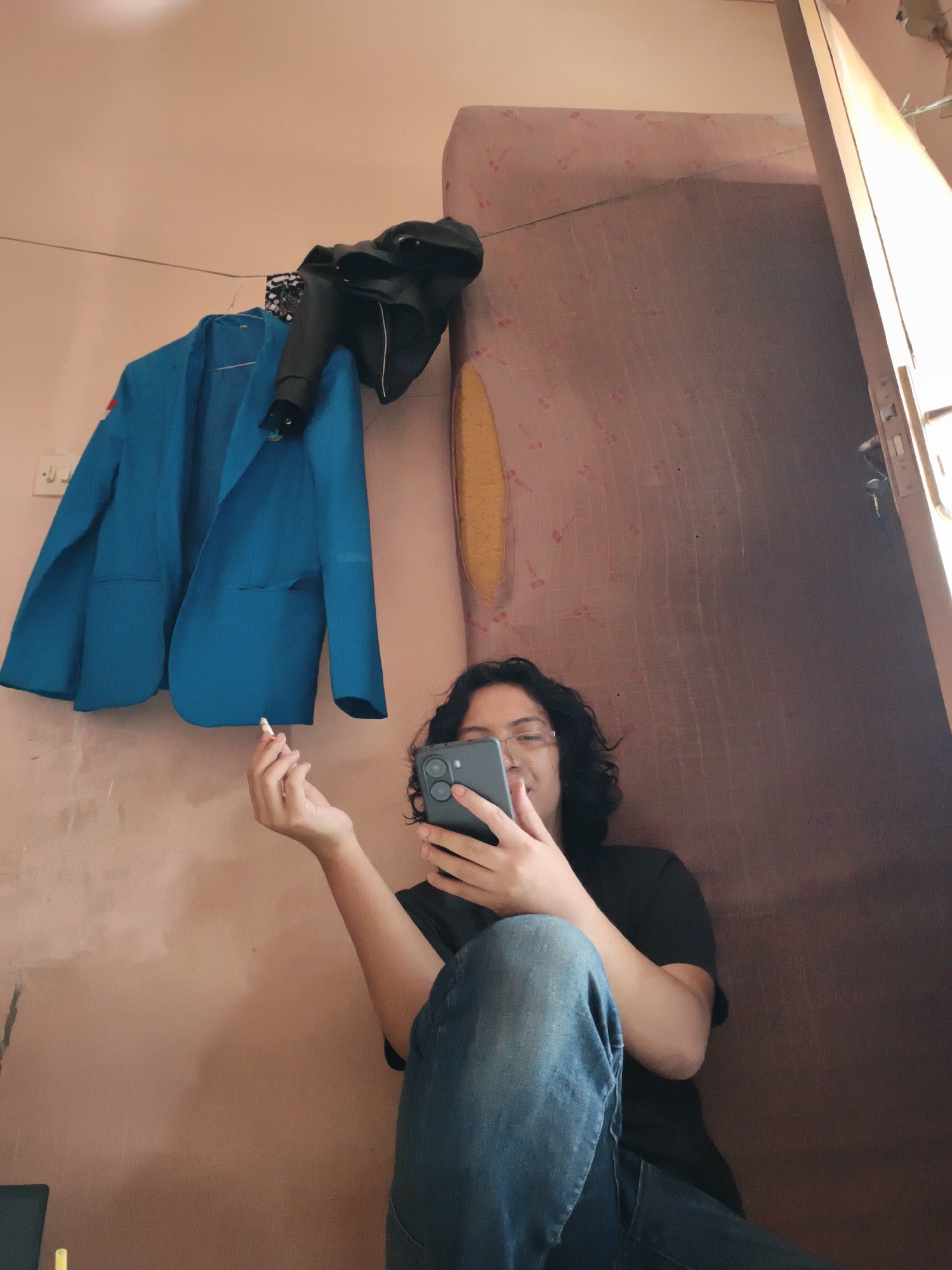 A person with dark, wavy hair and glasses is seated indoors, cross-legged, wearing jeans and a dark t-shirt. They are holding a smartphone in their hands and looking at the screen. In their left hand, they are holding what appears to be a cigarette. The background consists of a pink wall, a doorframe partially visible on the right, and items hanging on the wall to the left: a blue blazer and a black leather jacket. The location is in Cianjur, Indonesia.