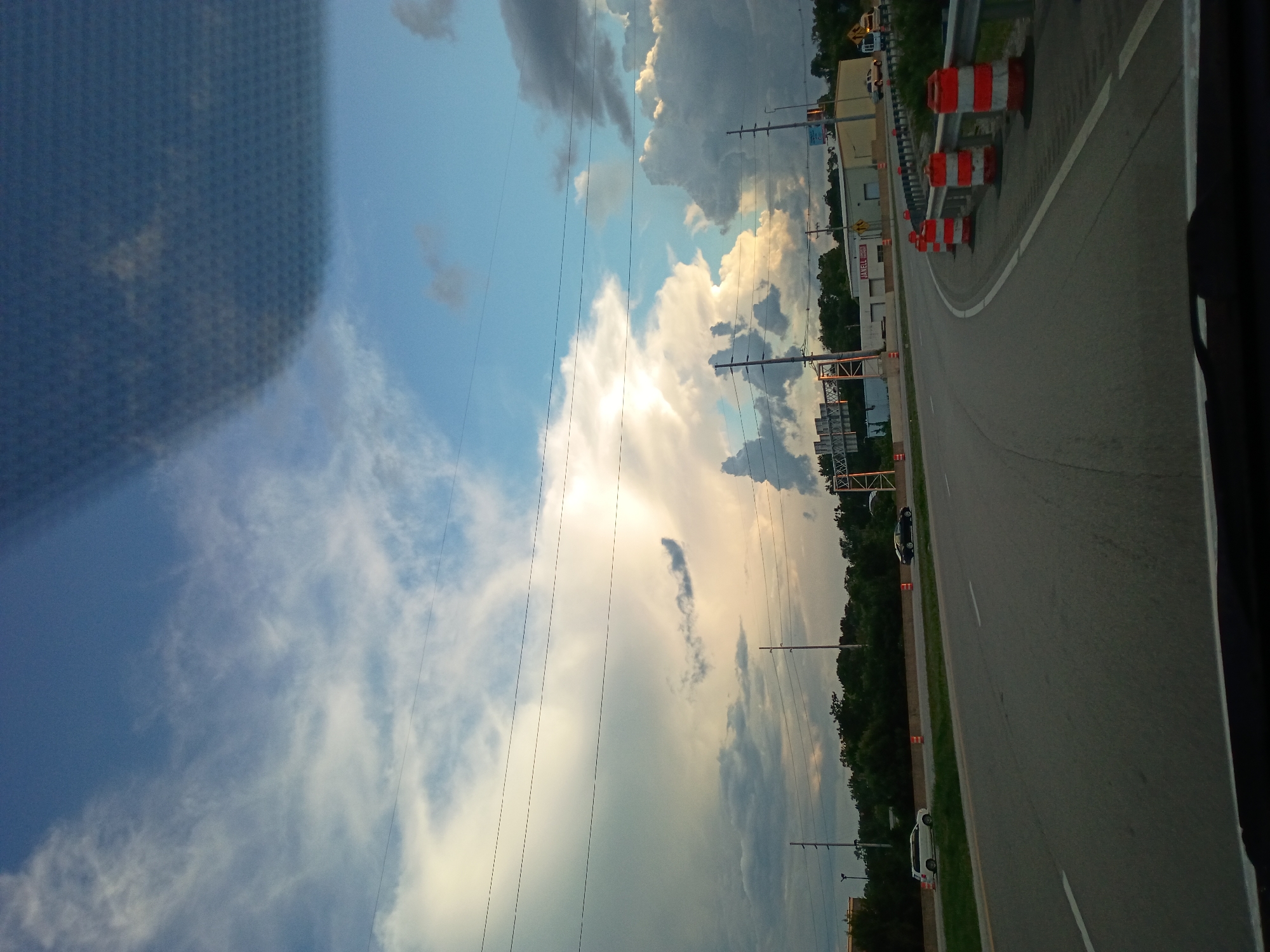 The image shows a view from a vehicle driving on a highway in Lexington, Kentucky. The highway has multiple lanes, and there are several traffic barrels lining the right side of the road, indicating construction or lane closures. In the distance, there are buildings, trees, and overhead power lines. The sky is filled with dramatic clouds, with some areas of bright sunlight breaking through. Two cars are visible on the highway, one white SUV and one black sedan. The overall scene suggests an outdoor environment, likely during the late afternoon or early evening, with a mix of natural and man-made elements.