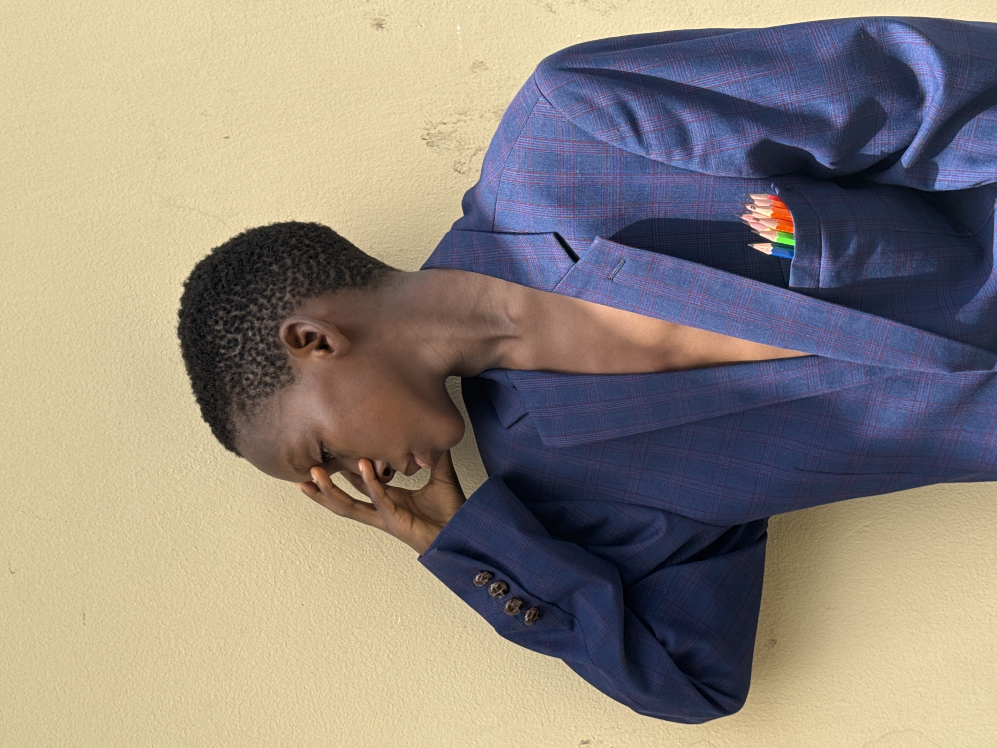 The image shows a dark-skinned person wearing a blue plaid suit jacket, with the jacket resting on a textured yellow surface, perhaps a wall. The person's head is turned and tilted downward, with their hand partially covering their face. A set of colored pencils is visible in the jacket's breast pocket. The location is Ado, Nigeria is not discernible from the image alone.