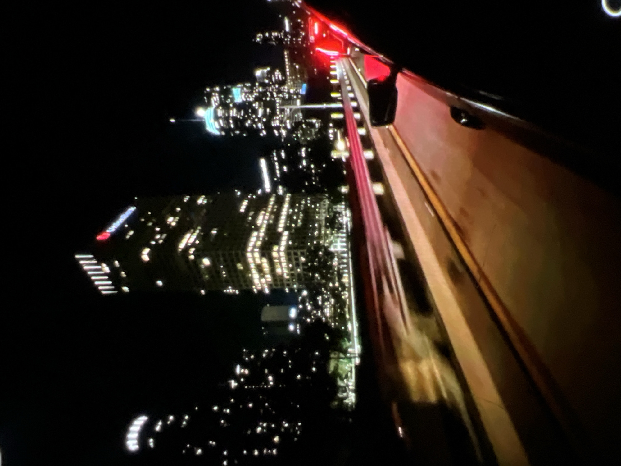 The image captures a nighttime view of a city skyline, likely taken from a moving vehicle. In the foreground, the right side of a car's interior is visible, including a side mirror and a portion of the car's exterior painted in red. The road ahead is illuminated by car headlights and streetlights, with streaks of red light indicating motion.
Several tall buildings dominate the background, with many windows lit up, suggesting a business district. The buildings are of varying heights and architectural styles. Some buildings are partially obscured by trees or other structures. The overall atmosphere is urban and vibrant due to the city lights.
Without specific landmarks or contextual information within the image, it is impossible to definitively identify the city as Lagos, Nigeria, or any other specific location. The image displays generic characteristics of a modern city at night.
