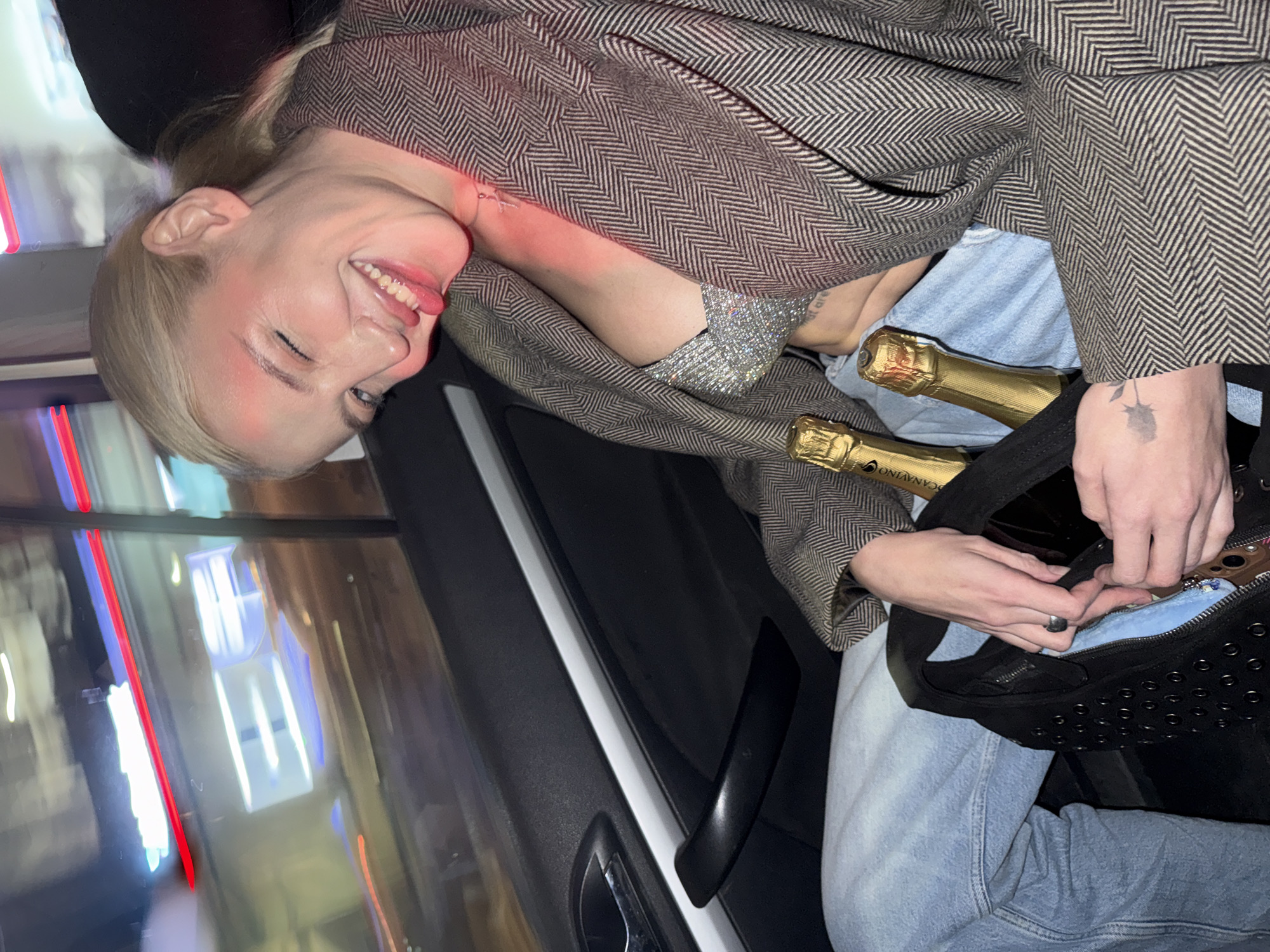 A woman with blonde hair is inside a vehicle holding a black studded purse in her lap with two bottles of champagne inside. She is wearing a gray jacket and blue jeans. The scene is likely in Belgrade, Serbia, with lights from the city visible through the car window.