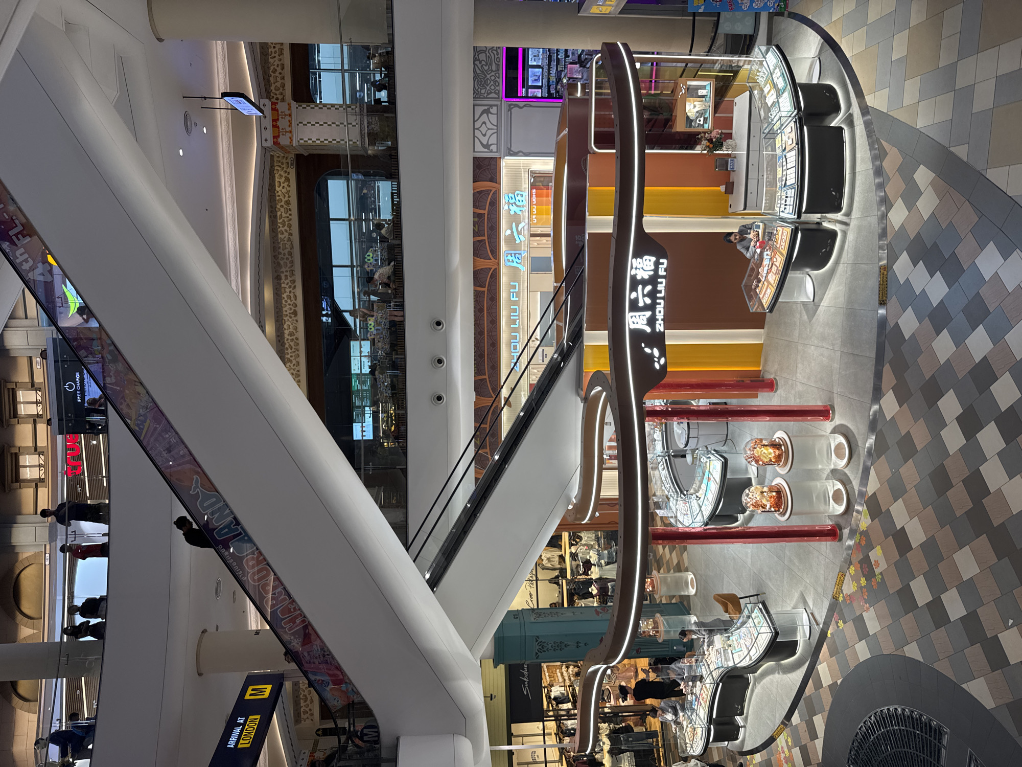 This is an elevated, wide-angle, interior shot of a busy shopping mall, likely in Pattaya City, Thailand, given the context. The image is dominated by a large, curved escalator system ascending diagonally through the frame. Below, a jewelry store named "ZHOU LIU FU" is prominently displayed with its illuminated signage and display cases filled with merchandise. Several customers are browsing the jewelry, and staff members are attending to them. The mall's architecture features multiple levels with shops and escalators visible in the background. The flooring is a geometric pattern of tiles in various shades of grey, brown, and beige. The lighting is a mix of ambient mall illumination and the bright lights of the stores. The overall impression is one of a modern, well-lit, and active retail environment.