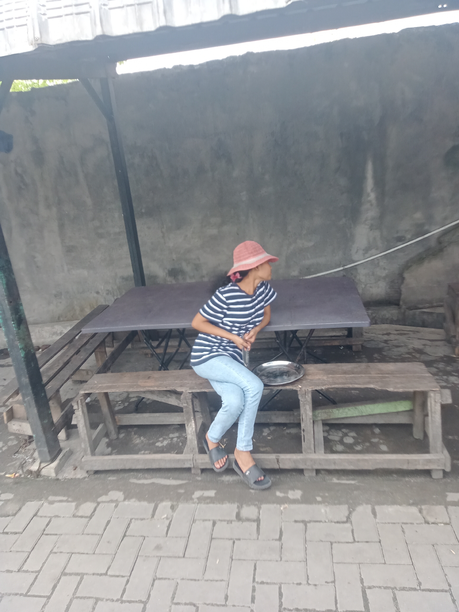 In Sunggal, Indonesia, a young female wearing a pink hat, a blue and white striped t-shirt, and light blue jeans is sitting on a wooden bench. She is looking to her right, away from the camera. A metal plate rests on the bench in front of her. A dark grey table is positioned behind the bench. The setting appears to be an outdoor covered area with a concrete wall and a corrugated metal roof. The ground is paved with interlocking bricks.
