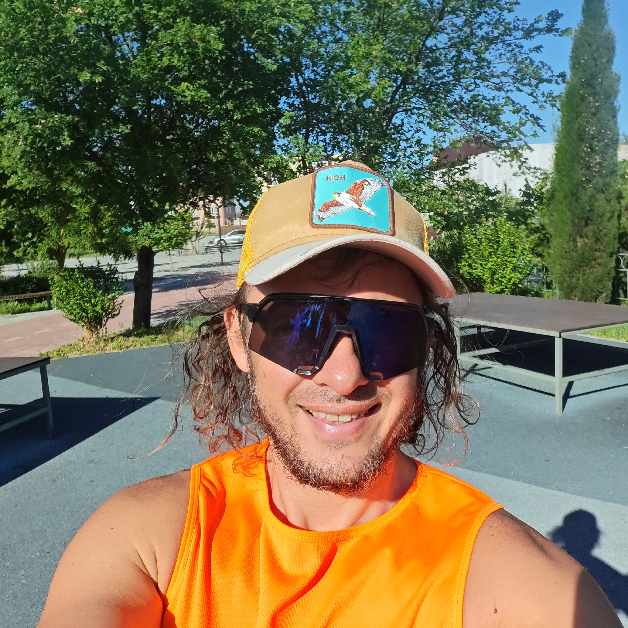 A selfie of a man wearing sunglasses and a hat with a bird patch and the word "HIGH" on it. He is also wearing an orange tank top. He is smiling. The background includes trees, a bench, and a building. The photo was taken in городское поселение Щелкино, Ukraine, as suggested by the available geotag data.