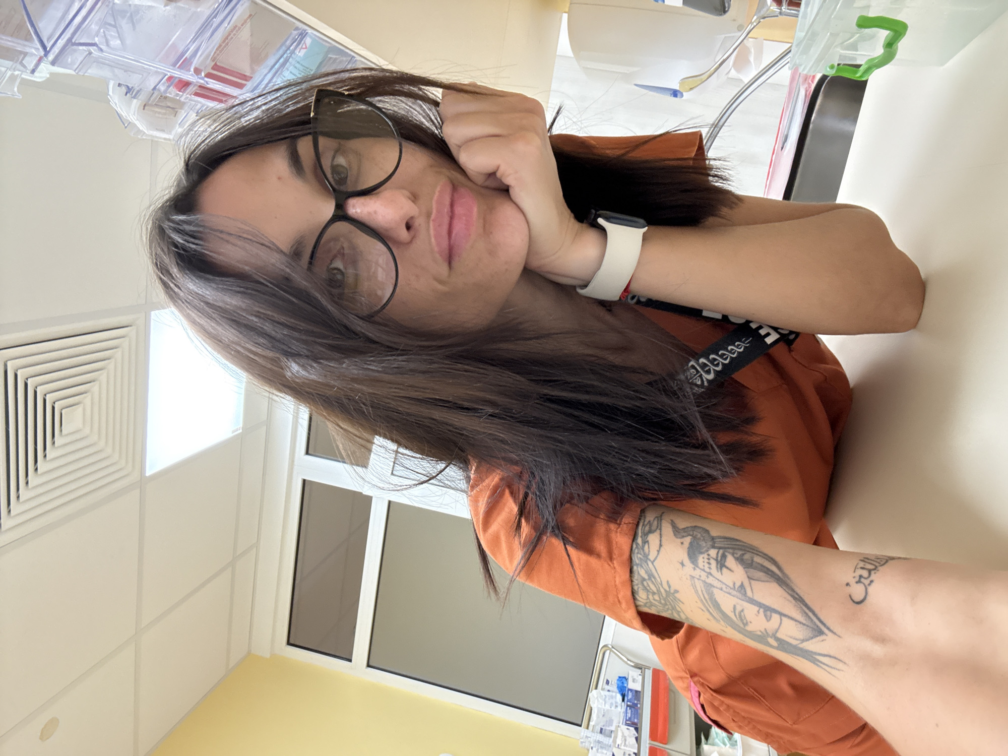 This image, taken in Pleven, Bulgaria, depicts a woman in a medical or laboratory setting, captured in a low-angle selfie. The woman has long, dark brown hair, wears dark-rimmed glasses, and has visible freckles. She is wearing an orange scrub top. Her left arm rests on a white counter, with her left hand supporting her chin. On her left forearm, there are two tattoos: one depicting two split faces, one with a horn, and another beneath it with Arabic script translating to "the religion" or "the faith." A white smartwatch is visible on her left wrist. The background features white grid ceiling tiles, a square air vent, and a rectangular fluorescent light fixture. A yellow wall section is visible on the left, next to a window with a dark frame. Various medical supplies, including boxes and a mobile cart, are present. On the right side of the frame, a clear, multi-compartment storage unit is mounted above the counter, and a clear plastic container with a green handle, along with a metallic sink faucet, is visible on the counter.
