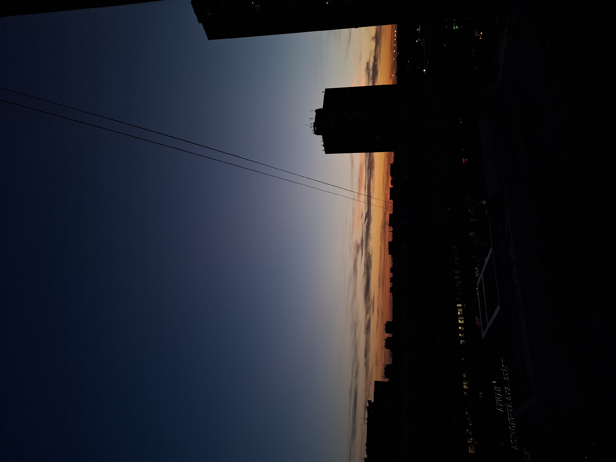The image depicts a cityscape at dusk, likely in Novi Sad, Serbia. The sky transitions from a dark blue to orange and peach hues near the horizon. Silhouettes of buildings, including high-rises, are visible against the sky. Two wires stretch across the sky, extending from the upper left to the center of the image. The lower right portion of the image is mostly dark, showing a structure with text obscured by shadows. No people are visible.
