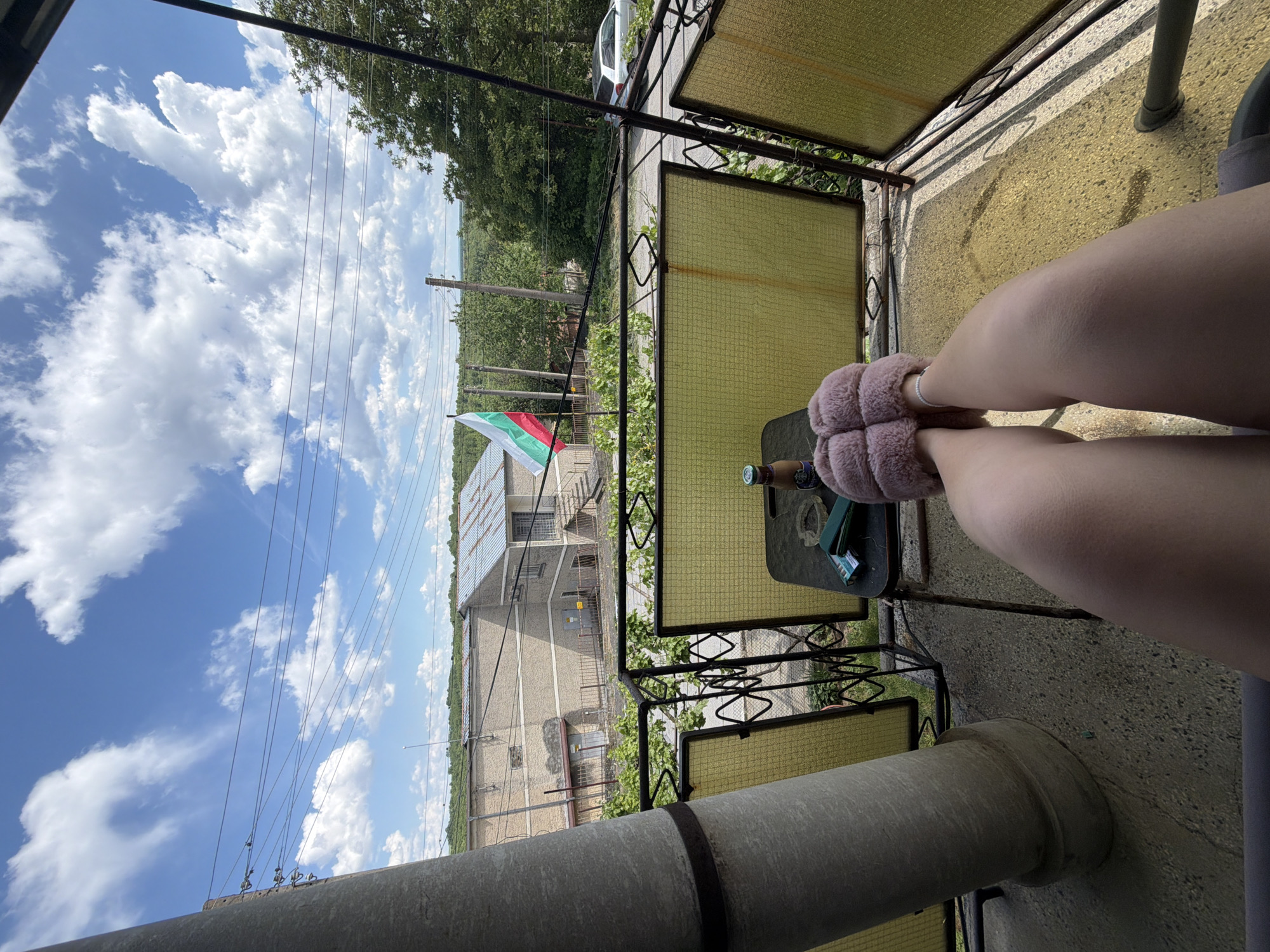 View from a balcony in Senovo, Bulgaria. Pink slippers are visible, along with a small table and a Bulgarian flag on a nearby building. Overhead power lines are visible against a blue sky with scattered clouds.