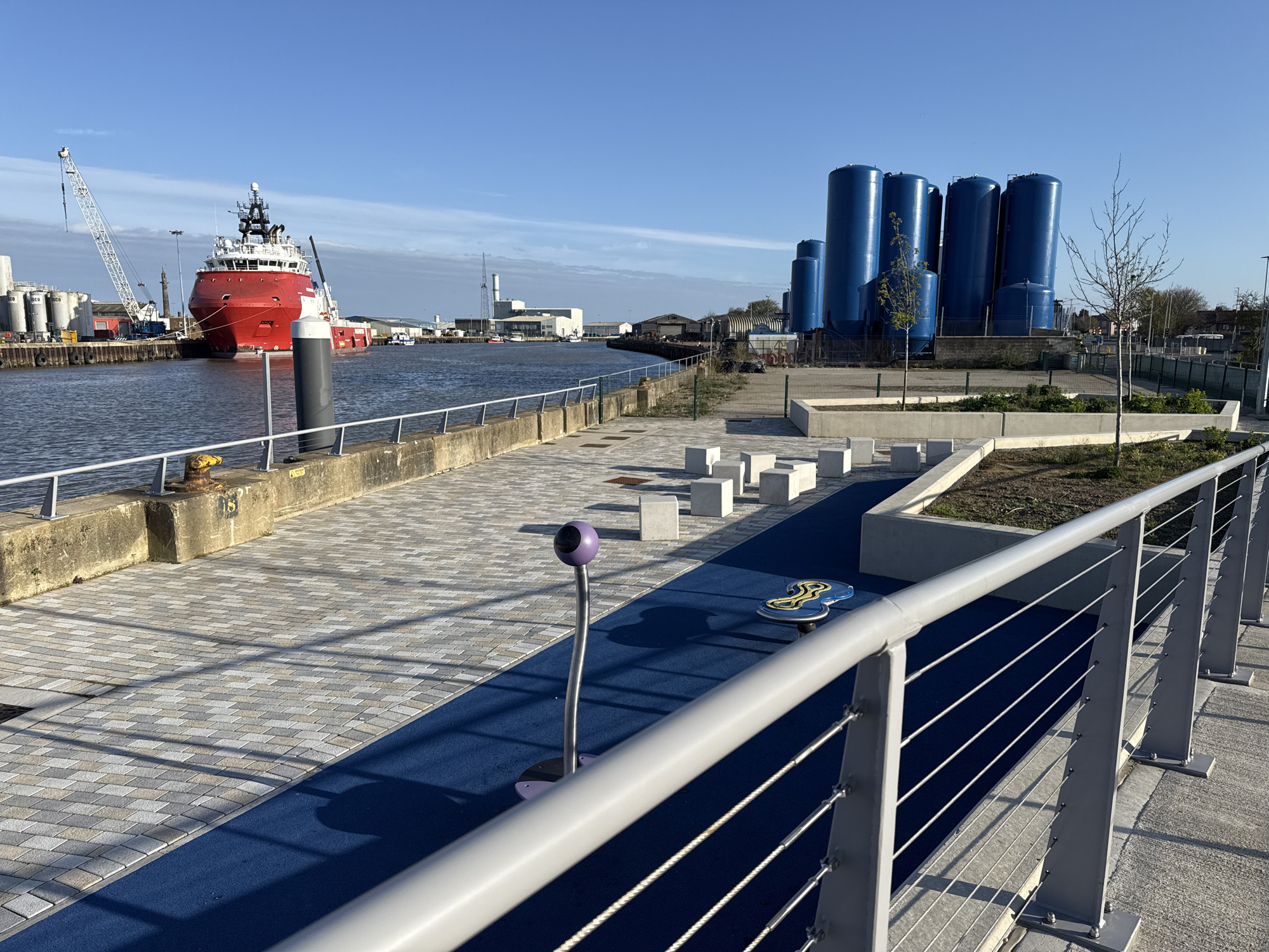 The image depicts a waterfront walkway in Gorleston-on-Sea, United Kingdom, featuring a paved area with a blue artificial surface and interactive playground elements, including square concrete blocks, a purple standing microphone device, and a blue spinning object. A modern metal railing runs along the right side of the walkway. Beyond the walkway, there is a concrete dock with a red offshore supply vessel docked along with large white tanks and a crane. In the background, there are a series of tall blue storage tanks. The sky is clear and blue.