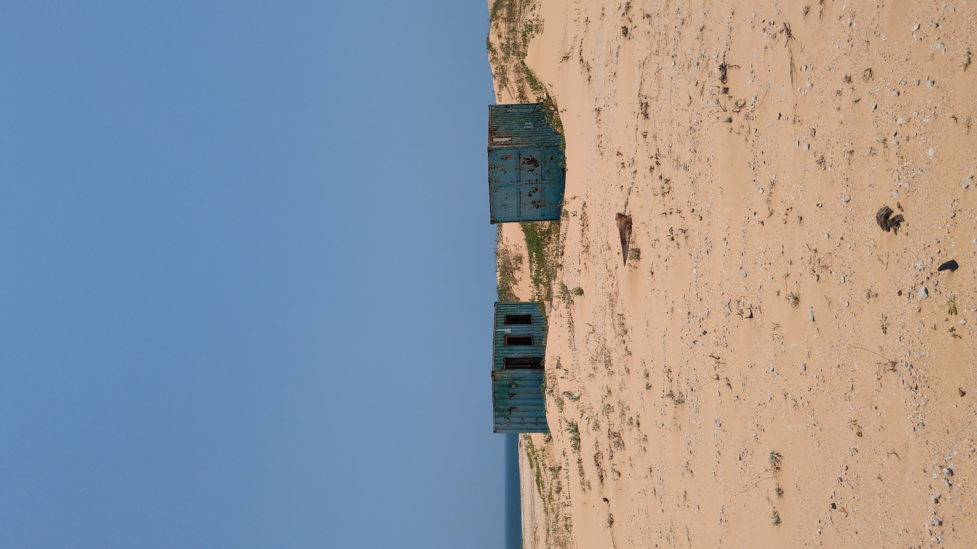 The image shows a sandy beach with two stacked blue shipping containers in Phuoc Dinh, Vietnam. The containers are rusty and appear abandoned. The sky is clear blue. The beach is sparsely vegetated.