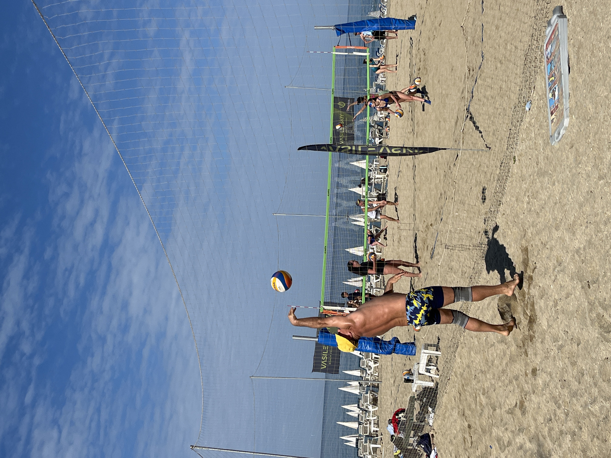 The image shows a beach volleyball game in progress on a sandy beach under a bright blue sky with scattered clouds. The beach has lounge chairs and umbrellas in the background, suggesting a resort or recreational area. A volleyball net is visible in the foreground and middle ground, with a man in blue and yellow patterned shorts about to hit a volleyball. He is wearing knee sleeves and is barefoot. Other people are playing volleyball on a nearby court. A sign with "VASILE BEACH HOUSE" is positioned in the middle ground. A large advertisement billboard is visible to the right side of the frame. The scene is taking place in Alanya, Turkey.