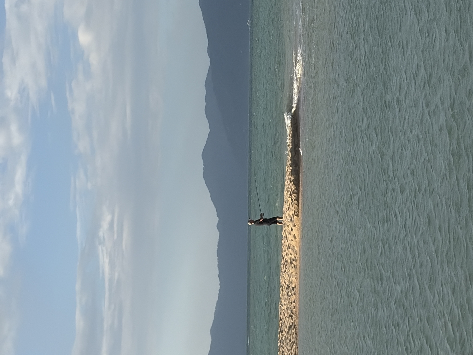 A person is fishing on a narrow strip of sand that juts out into a calm, greenish-blue body of water. The person, dressed in dark clothing, stands with a fishing rod, casting into the water. The strip of land is covered in sand and pebbles, with a small wave breaking on its edge. In the background, a range of dark, hazy mountains stretches across the horizon, suggesting a coastal or island location. The sky above is a pale blue with scattered white clouds, indicating fair weather. The scene appears to be set during the daytime, likely late morning or early afternoon, given the brightness of the sky. The overall mood is one of peaceful solitude and recreation. The location is identified as Ko Samui District, Thailand. There is no visible text in the image.