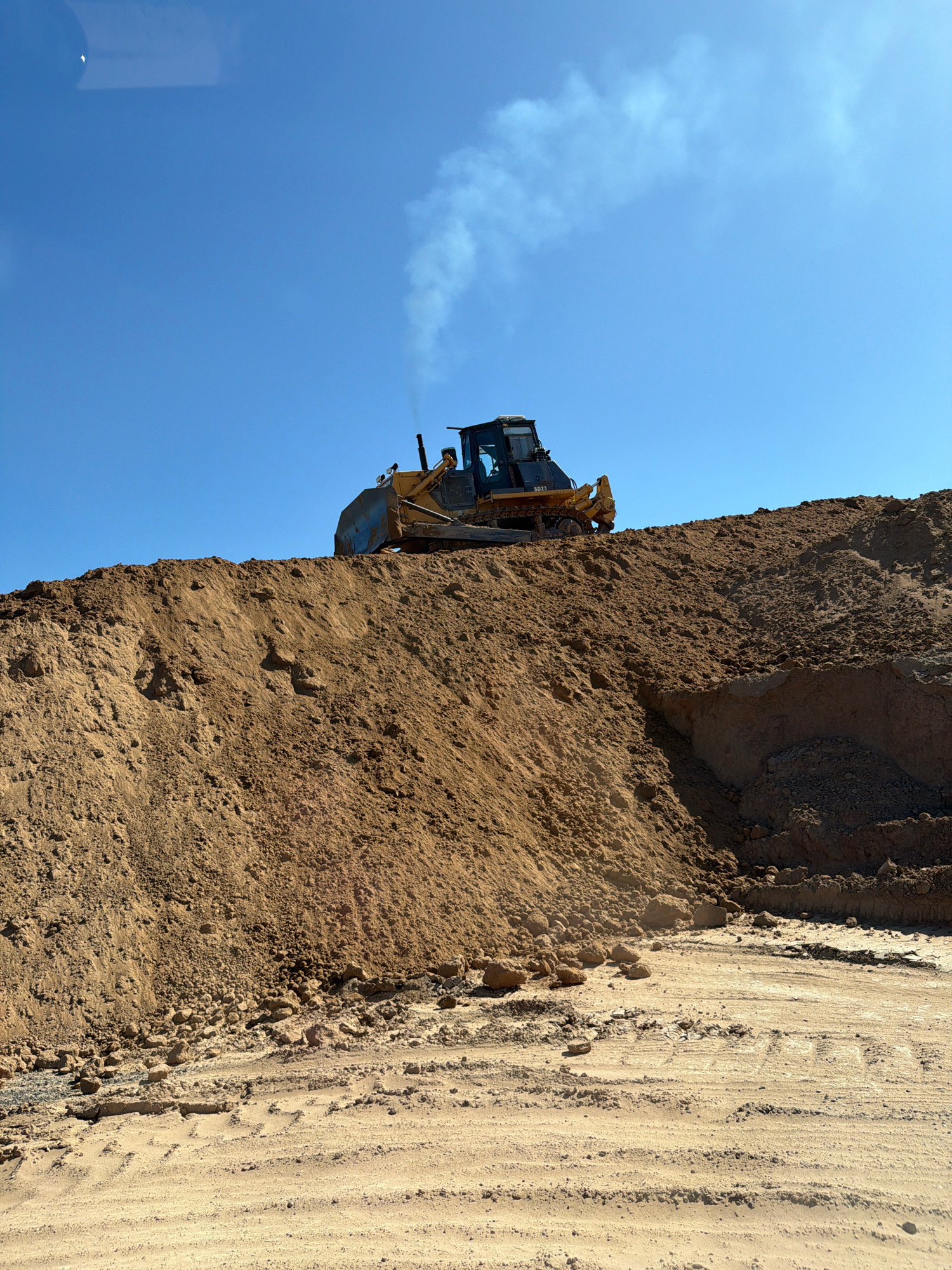 An outdoor, medium-shot photograph captures a yellow bulldozer with black accents operating on a large dirt mound under a clear blue sky. The bulldozer is positioned on the crest of the mound, facing left, and appears to be pushing dirt. A plume of white smoke emanates from its exhaust pipe. The mound itself is composed of light brown, granular soil, with some scattered rocks and debris visible on its surface and at its base. The foreground shows a flattened dirt area with tire tracks, indicating recent activity. The overall scene suggests a construction or earthmoving site.