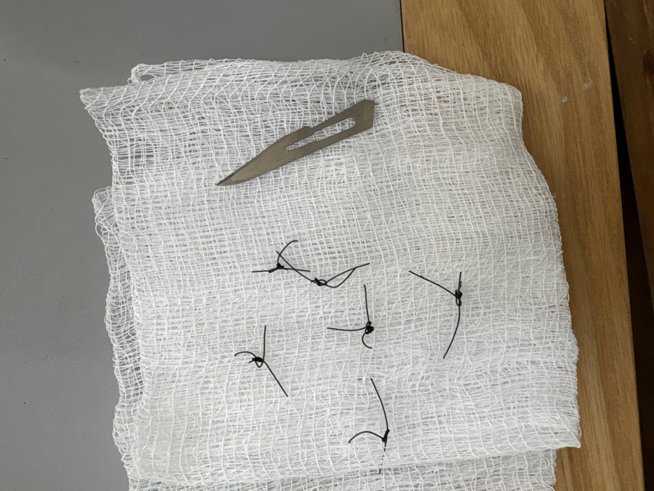 A metal scalpel blade rests on a white gauze pad, which is also scattered with short lengths of black surgical suture. The pad sits partially on a grey surface and partially on a light brown wood surface. The sutures are tied in knots at their midpoints, with loose ends extending outwards. The gauze's woven texture is visible.