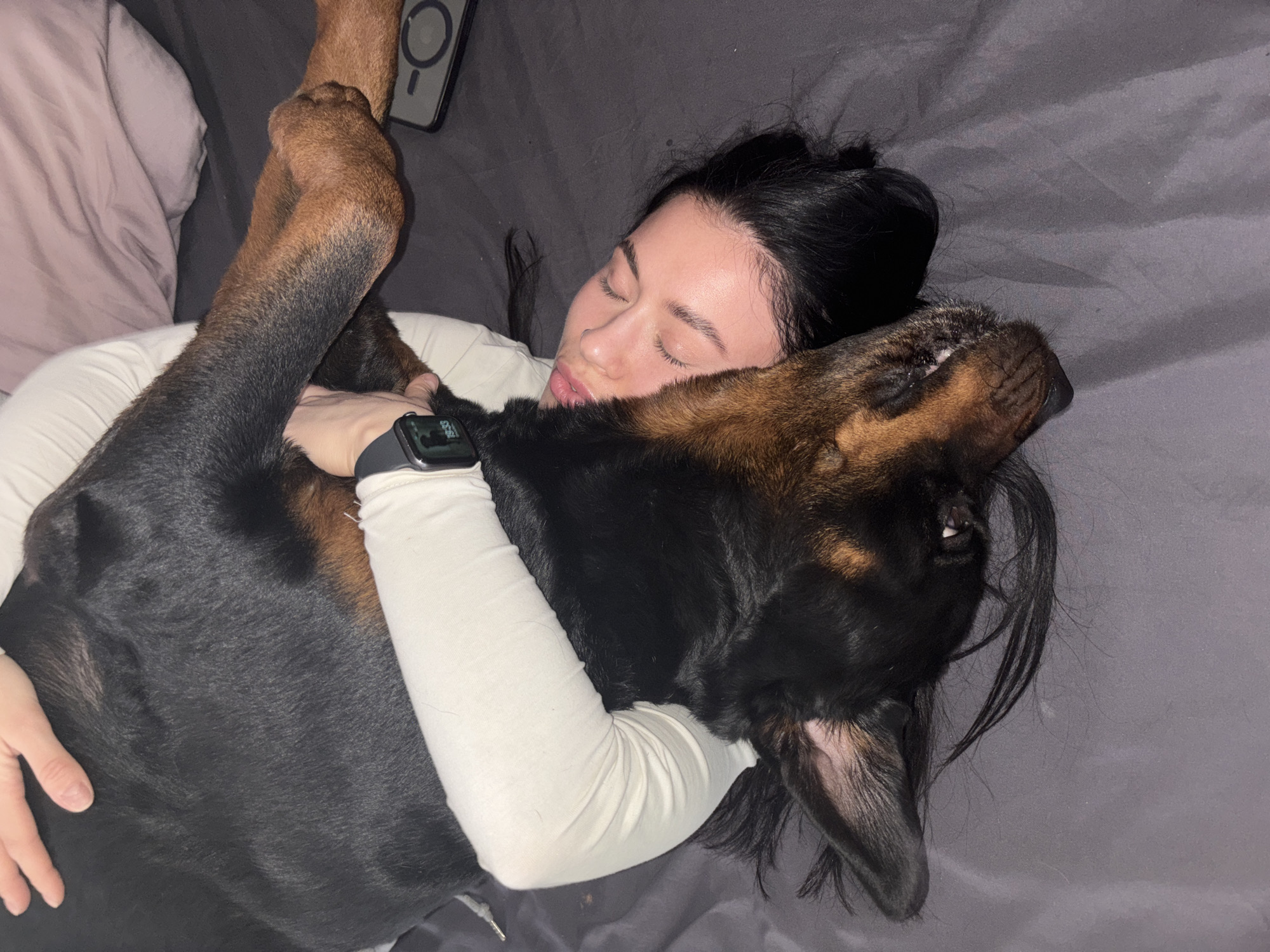 A person is lying in bed with a large dog. The person, likely female, has dark hair and is wearing a long-sleeved white shirt. Her eyes are closed and she is turned towards the dog, appearing to be embracing it. The dog is mostly black with brown markings on its face and legs. The dog's head rests near the person's face. The person is wearing a smartwatch on their left wrist. A smartphone is lying near the head of the bed. The bed has grey bedding. The location of Perm, Russia, is not discernible from the image.
