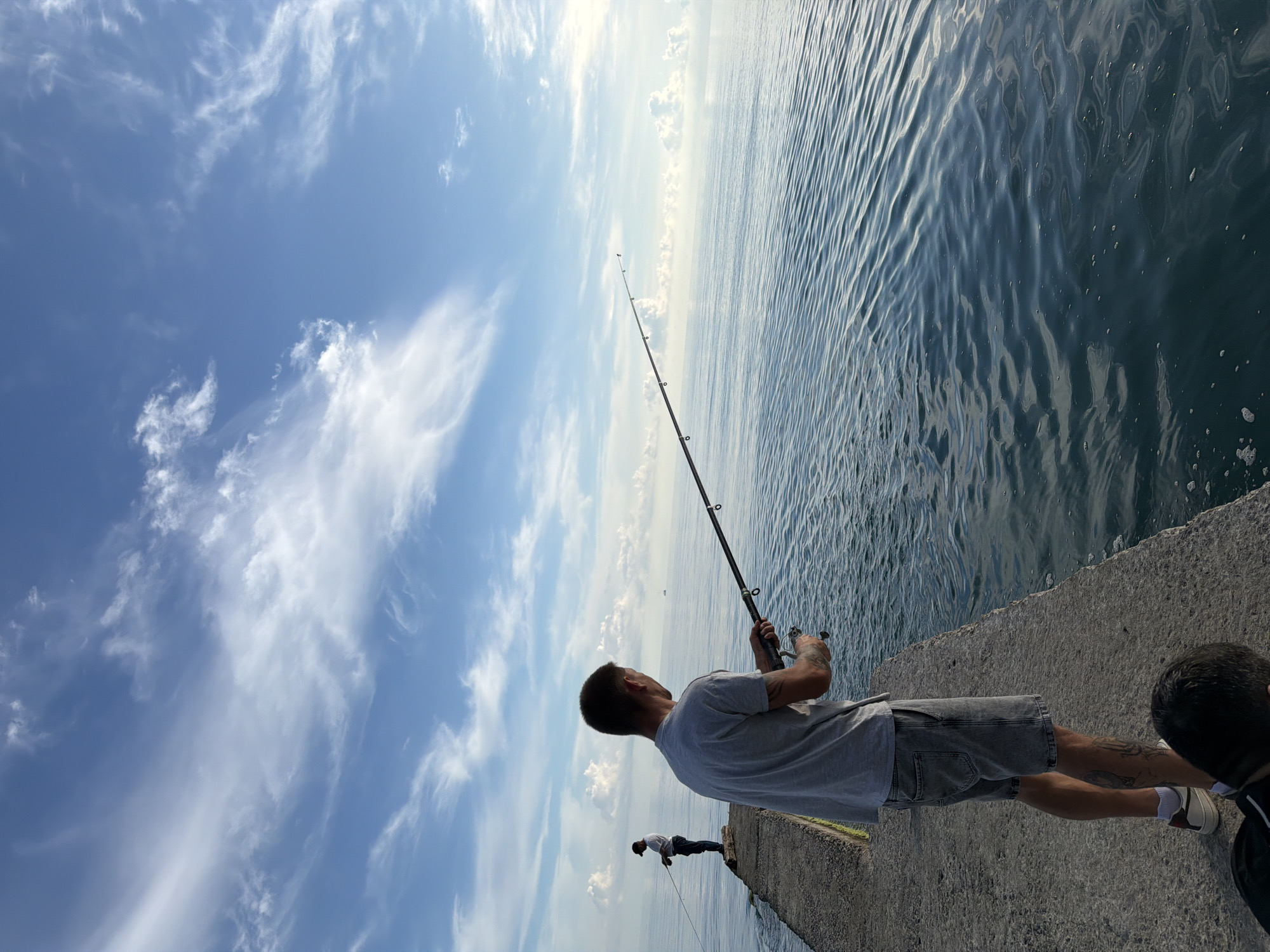 In Dederkoy, Russia, on a concrete pier extending into a calm sea under a bright blue sky with scattered white clouds, two individuals are engaged in fishing. The foreground shows a man facing away from the camera, holding a fishing rod with a reel, actively fishing. He is wearing a grey t-shirt, denim shorts, and sneakers. His tattoo is visible on his left leg. Further down the pier, a second person, also facing away, is standing and fishing with a rod. The sea is calm with gentle ripples, and the horizon is visible in the distance.