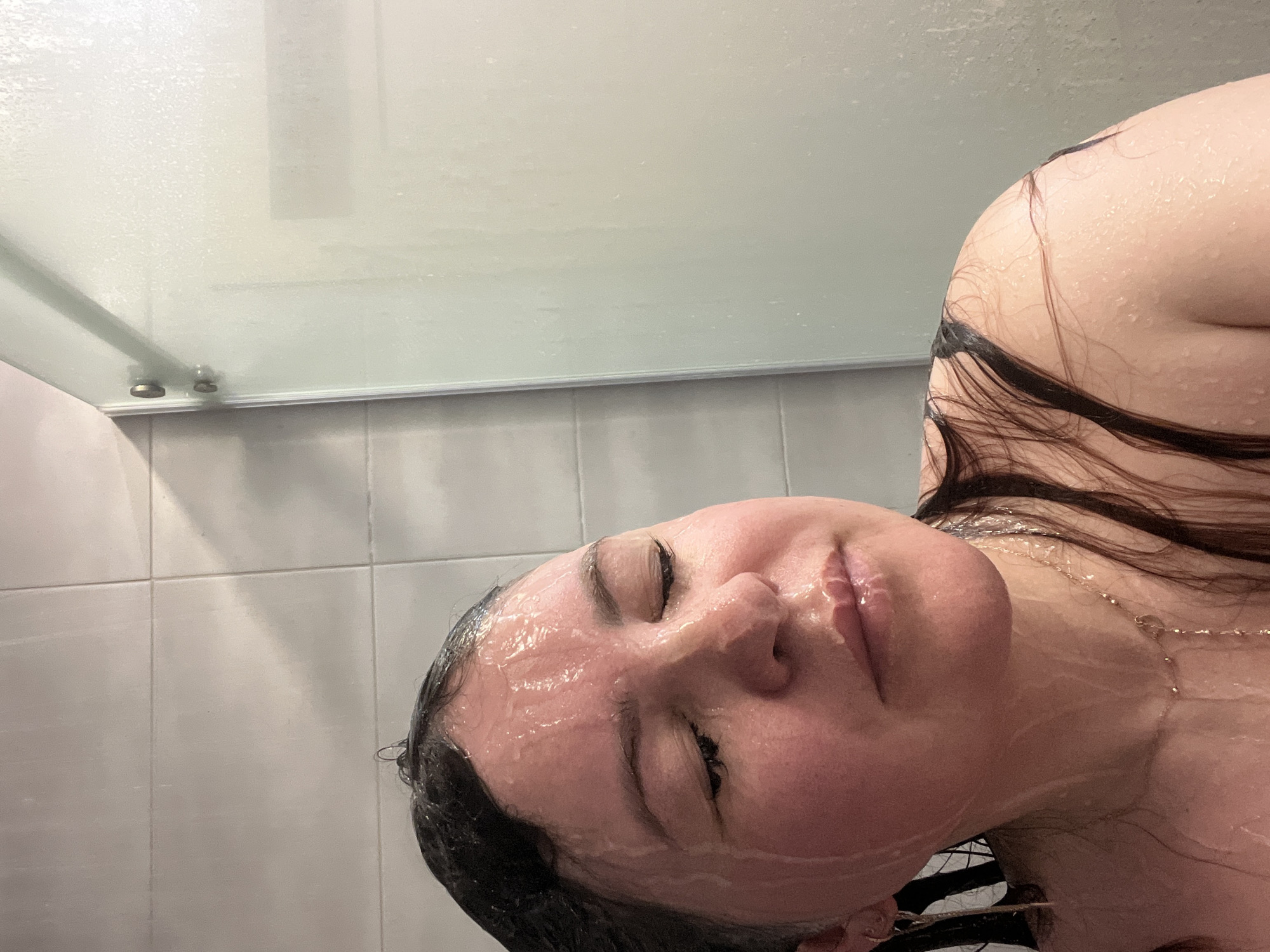 A woman is in a shower with water running over her face and body. The shower has white tile walls and a glass door. The location is not discernible as Noviy, Russia.