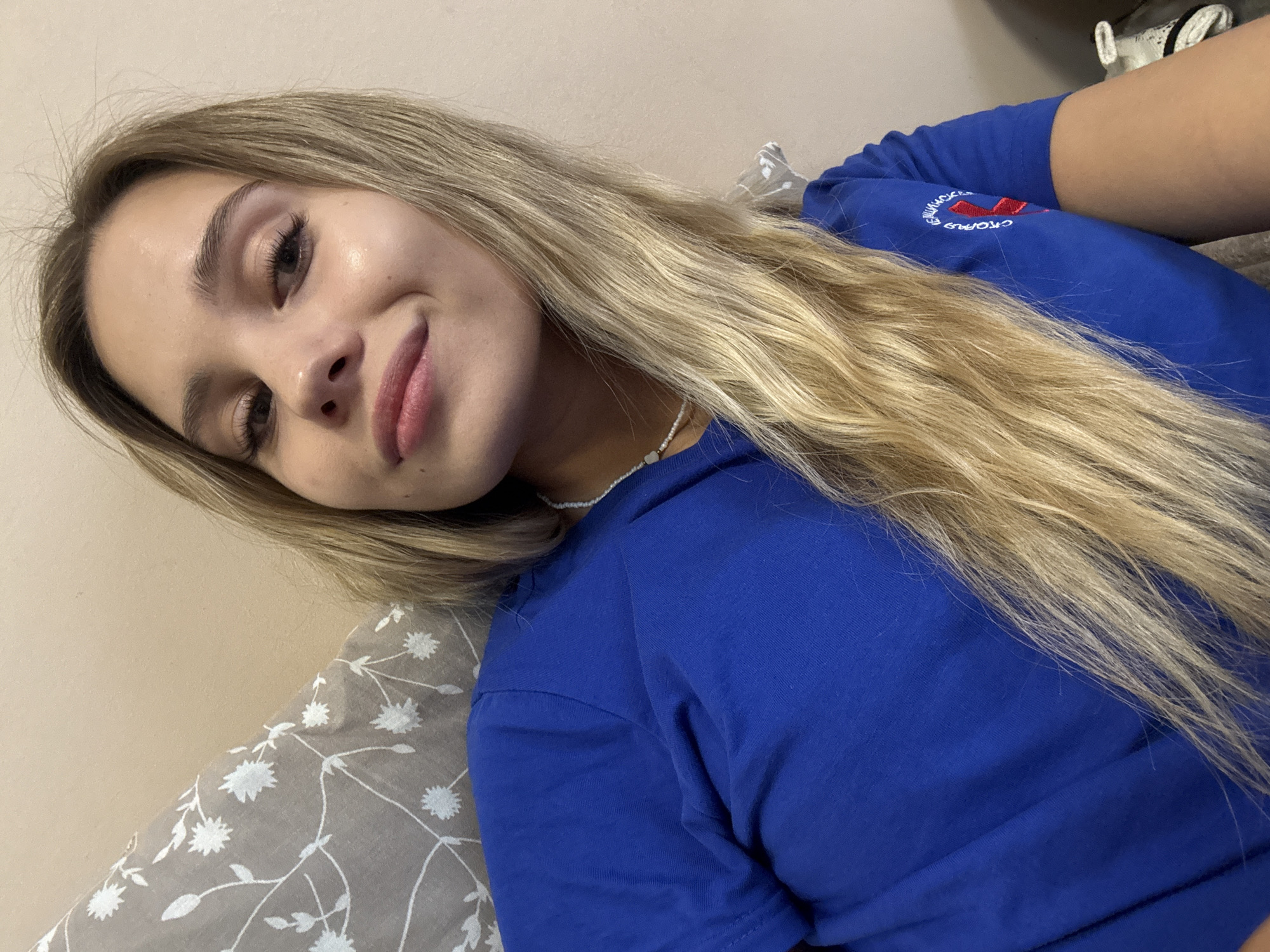 A young woman with long, blonde hair and a slight smile is depicted in a close-up, upward-angled shot, likely a selfie. She is wearing a royal blue short-sleeved t-shirt and a delicate white beaded necklace with a small, light-colored charm. The left sleeve of her t-shirt features white embroidery with Cyrillic text, partially legible as "СТОРОНА" (Side/Party), followed by additional unreadable Cyrillic script and a distinct red ribbon symbol. The background consists of a light beige wall on the right and top, and a gray fabric with a white floral or botanical pattern, likely a pillow or blanket, on the left and behind her shoulder. The scene is located in the city of Chaikovsky, Russia.