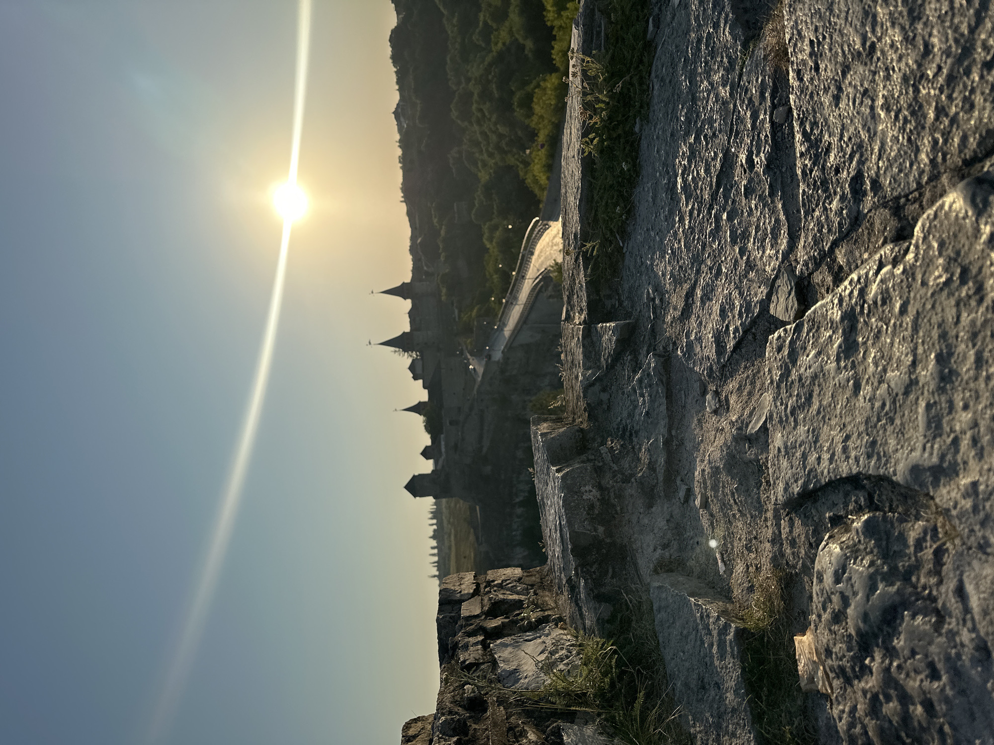 This is a vertical, eye-level shot of a sunlit stone embankment overlooking a canyon with a castle and a bridge. The sun is low in the sky, casting a golden light on the scene and creating a lens flare in the upper left portion of the frame. In the foreground, large, rough-hewn stones make up the embankment, with sparse patches of dry grass and small plants growing in crevices. The middle ground features a deep canyon with a winding road and a bridge crossing it. Beyond the canyon, the Kamianets-Podilskyi Castle, with its distinctive towers and spires, is silhouetted against the bright sky. Lush green trees cover the slopes of the canyon, contrasting with the stone structures. The sky is a clear, pale blue, with a subtle cloud streak extending from the sun. The overall impression is one of historical grandeur bathed in the warm glow of sunset.
