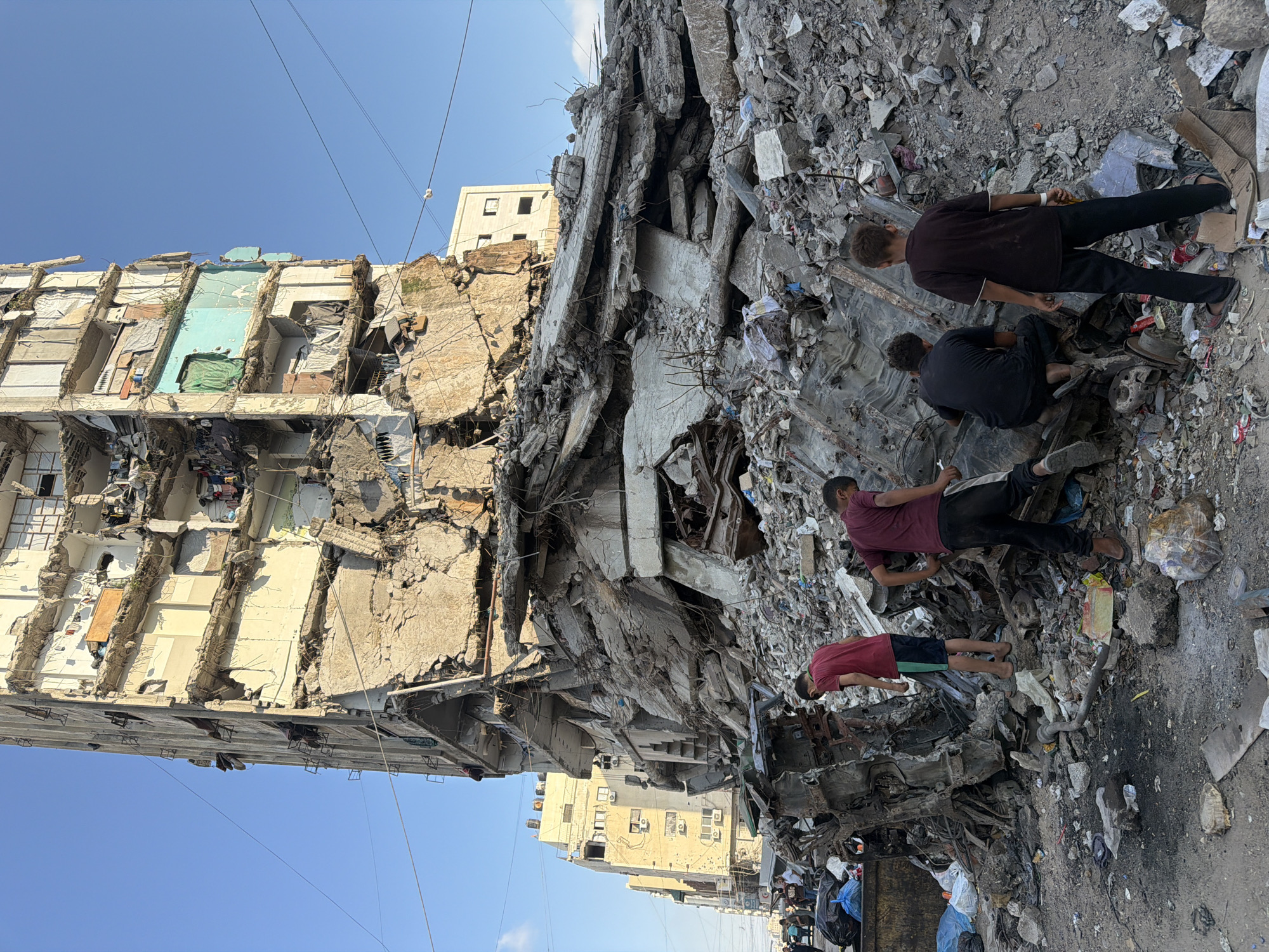 The image depicts the aftermath of an airstrike in Gaza City, Palestinian Territories, showing the severe damage to a residential building. A tall, multi-story apartment building is visibly fractured and partially collapsed, with concrete debris forming a large pile in the foreground. Several people, including children, are seen among the rubble, some appearing to be searching through the debris. The sky is clear and blue, contrasting with the destruction below. In the background, other buildings, some damaged and some appearing intact, line the street, alongside electrical wires and poles. The scene is one of devastation, with scattered debris and damaged structures indicating a recent destructive event.
