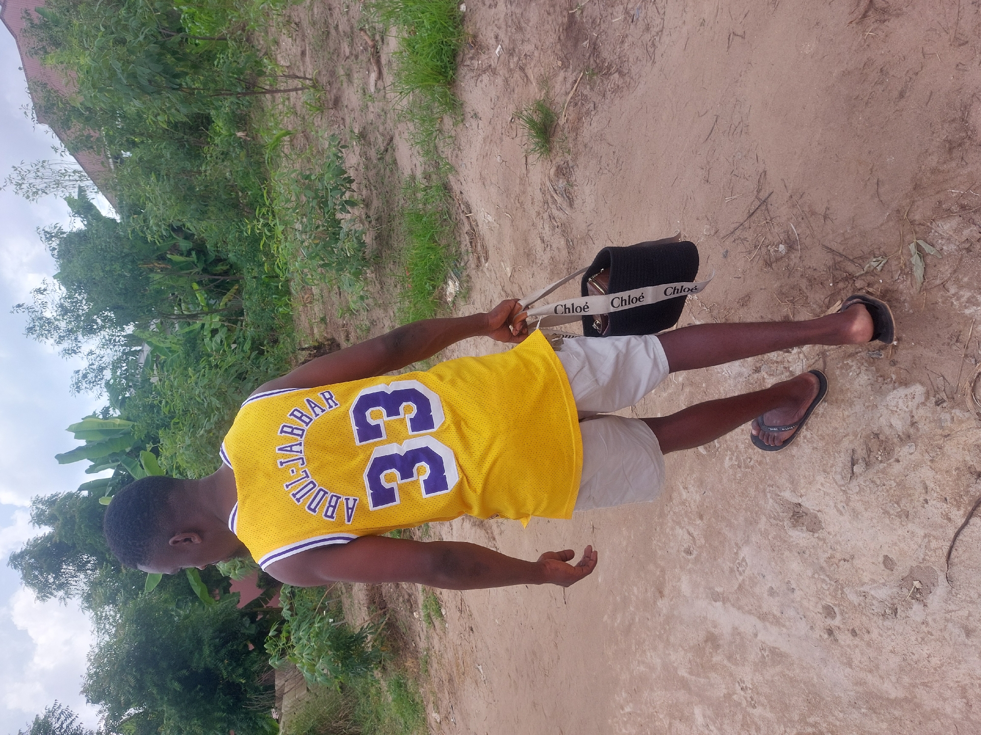 A man wearing a yellow Kareem Abdul-Jabbar Lakers jersey with the number 33 and gray shorts is seen from behind while standing on a sandy path. He is holding a black purse with white "Chloe" straps. He is wearing black sandals. The surrounding environment consists of foliage and trees. The location is likely Dar es-Salaam, Tanzania based on the user's prompt.