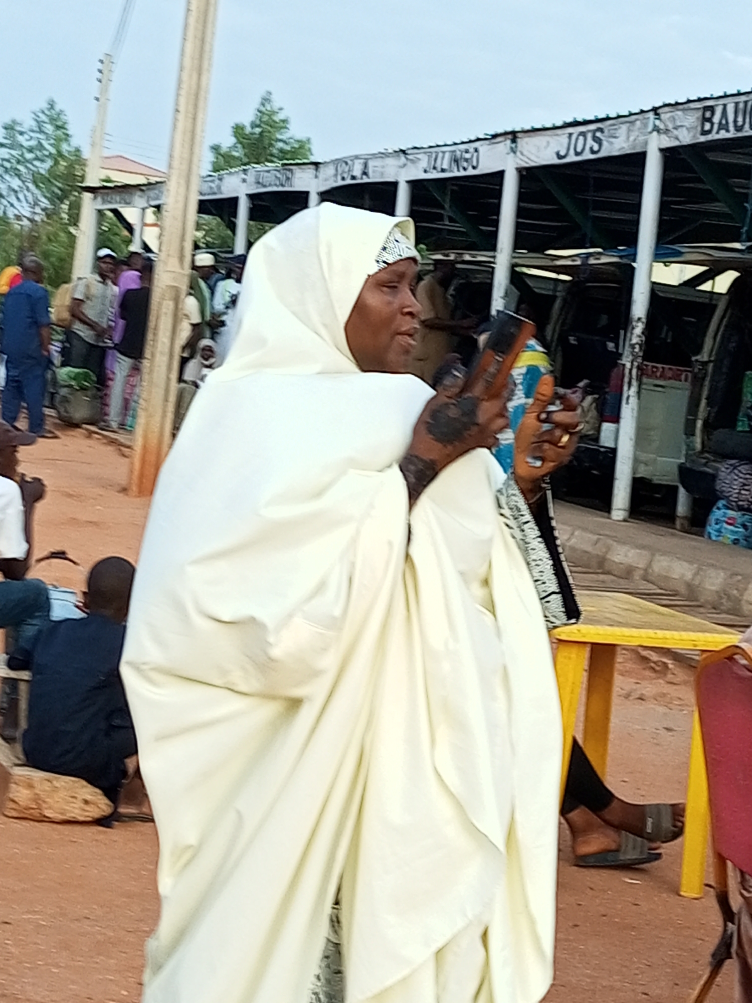 The image captures an outdoor scene, likely a busy market or transportation hub, in Gombe, Nigeria. A woman, wearing a white hijab and a flowing white garment, is in the foreground, holding a mobile phone. Her hands show intricate henna designs. Behind her, a diverse group of people are visible, some appearing to be passengers or vendors. The background features a covered structure with signage that reads "JALINGO JOS BAU". The ground is a sandy, reddish-brown color. Several vehicles, possibly minibusses, are parked under the covered area, suggesting a terminal or waiting area. The overall impression is one of a lively, everyday scene in a Nigerian city.