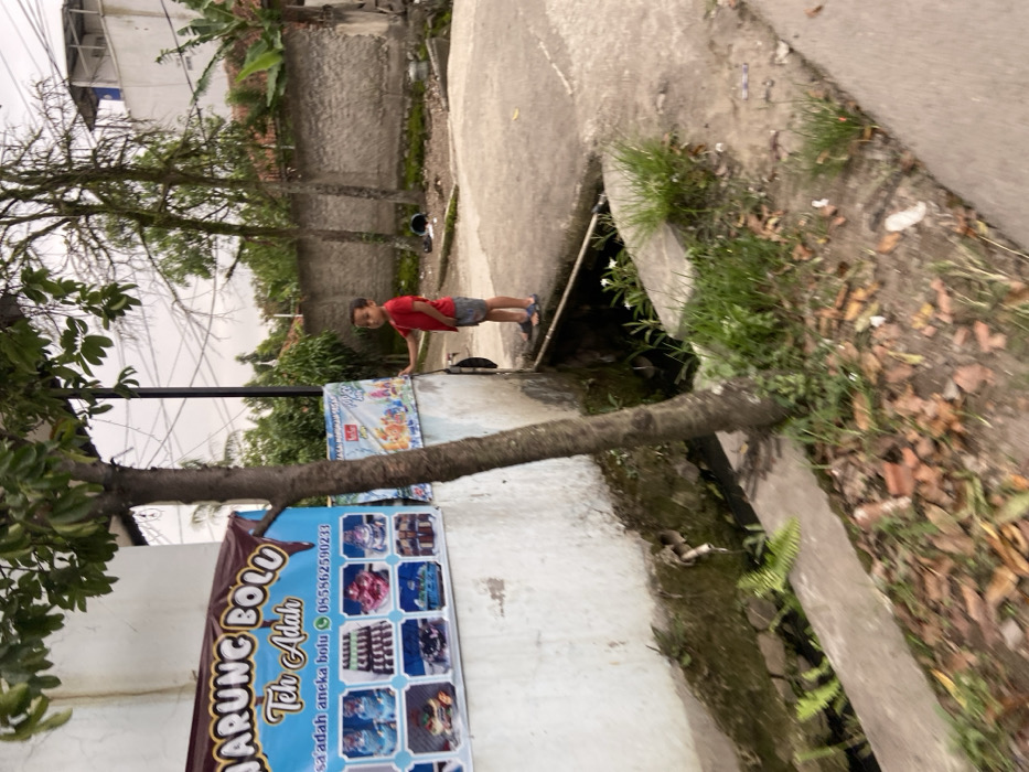 The image shows a young boy in a red shirt and blue shorts standing on a sidewalk in Margaasih, Indonesia. The boy is looking to the left, with his left hand on a wall. The wall is painted white and has a sign advertising a bakery called "Warung Bolu Teh Adah". The sign depicts various kinds of cakes and pastries. The boy is standing next to a large tree with a thick trunk. There is a small, open drainage ditch between the sidewalk and the wall. The ditch is filled with water and small plants. The ground around the ditch is covered in leaves and debris. There is a white building in the background, with other houses visible beyond it. The area is generally quite calm and serene, with few people or vehicles present. The image was taken in Margaasih, Indonesia at the coordinates -6.953 latitude and 107.551 longitude.