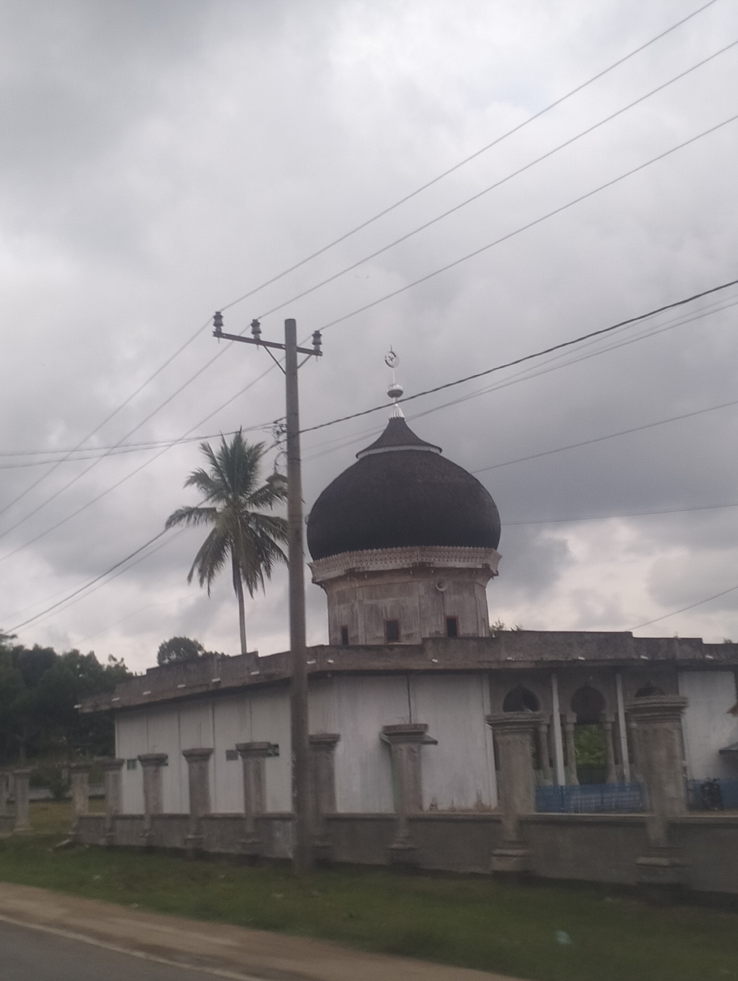 The image shows a mosque with a dark dome and a crescent moon finial, situated in Teupinreusep, Indonesia. The mosque has a white facade and a series of decorative pillars along its perimeter. A tall utility pole with overhead wires stands in front of the mosque, and a coconut palm tree is visible to the left of the pole. The sky is overcast with clouds. The image appears to be taken from a moving vehicle, indicated by the blurred foreground of a road.