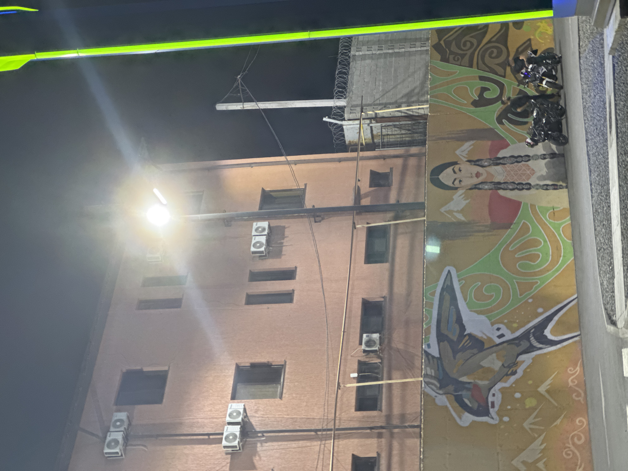 This media file captures a nighttime scene of an urban alleyway in Shymkent, Kazakhstan. The primary subjects are two motorcycles parked on a sidewalk that is adorned with a vibrant mural. The mural depicts a stylized representation of a woman with long braids and a bird in flight, set against an abstract, swirling background in earthy tones and greens. The motorcycles appear to be dark-colored and are positioned in front of the mural.
The setting is a narrow street or alleyway adjacent to a multi-story building. The building's facade is made of light brown bricks and features several windows and air conditioning units mounted on the exterior walls. A bright light fixture illuminates the scene, casting a strong glow and creating lens flare. Above the building, a metal structure with barbed wire is visible, suggesting a security measure or industrial element. In the upper portion of the image, a bright green, illuminated sign or architectural feature stretches across the frame.
The time of day is clearly night, indicated by the darkness of the sky and the artificial lighting. There are no clear indicators of weather, but the absence of precipitation is implied. The overall mood is somewhat mysterious and industrial, with the vibrant mural offering a splash of artistic expression against the utilitarian backdrop of the building and street. There is no visible text in the image. The presence of the motorcycles suggests that people may have recently been or are about to be present, but no individuals are directly depicted in the frame.