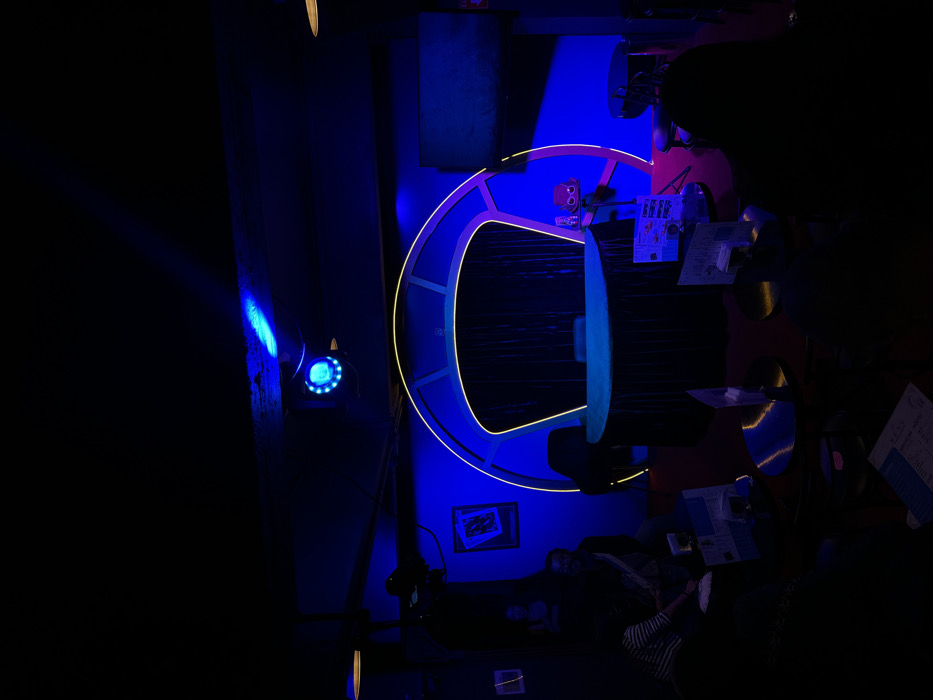 An interior view of a themed entertainment venue, likely a bar or lounge, located in Mexico City, Mexico, is depicted under deep blue ambient lighting. The central feature is a large, stage-like archway illuminated with yellow and purple neon or LED strips, designed to resemble a futuristic spaceship bridge. A semi-circular counter, draped in dark fabric, is positioned in front of the archway, with a dark curtain visible behind it.
To the left of the archway, a framed picture hangs on a blue-lit wall, and a spotlight projects blue light from the ceiling. Further left, three individuals are seated or standing in the dimly lit area: one person in a striped shirt is seated at a table examining what appears to be a menu, another person with a beard is partially visible behind them, and a third individual is indistinctly seen deeper in the background.
To the right of the archway, a flat-screen monitor is mounted on the wall. Below it, a small stand holds a red, stylized character figure resembling a "redshirt" from science fiction. In the foreground and midground, multiple round, dark tables are present. Some tables hold illuminated menus or information stands. One individual's back is visible in the lower right foreground, seated at a table, facing towards the central archway. The floor appears to be carpeted in a dark color. The overall atmosphere is dark and futuristic due to the prominent blue lighting and themed decor.