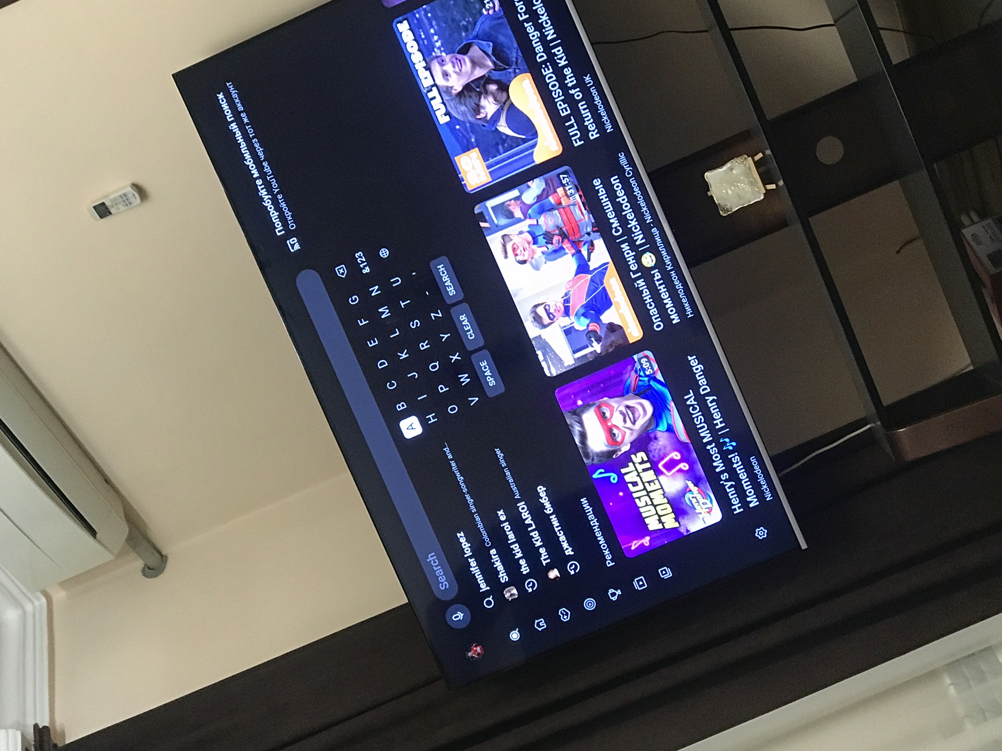 A television displaying a YouTube search interface with video suggestions, including content related to Nickelodeon's "Henry Danger," dominates the frame. The YouTube search bar is active with "Jennifer Lopez" as a recent query. The setting appears to be an indoor space, with a wall-mounted air conditioner visible on the left. The television is mounted above a dark-colored cabinet or shelving unit. A remote control is positioned on the wall above the television. The content is in English, with some Cyrillic characters. There is no information to support that the location is in Yerevan, Armenia.