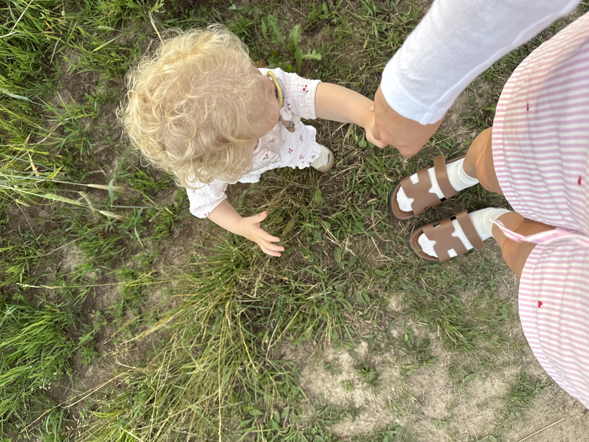 This is a first-person perspective, eye-level shot, taken outdoors on a dirt path with scattered grass. The image features a toddler with curly blonde hair wearing a white dress with a cherry print, white socks, and tan sandals. The toddler is holding the hand of an unseen person. In the foreground, the legs and feet of the unseen person are visible, wearing pink and white striped shorts and brown sandals with white socks. The scene appears to be in a rural or park-like setting. The location is not specified in the image or its metadata.