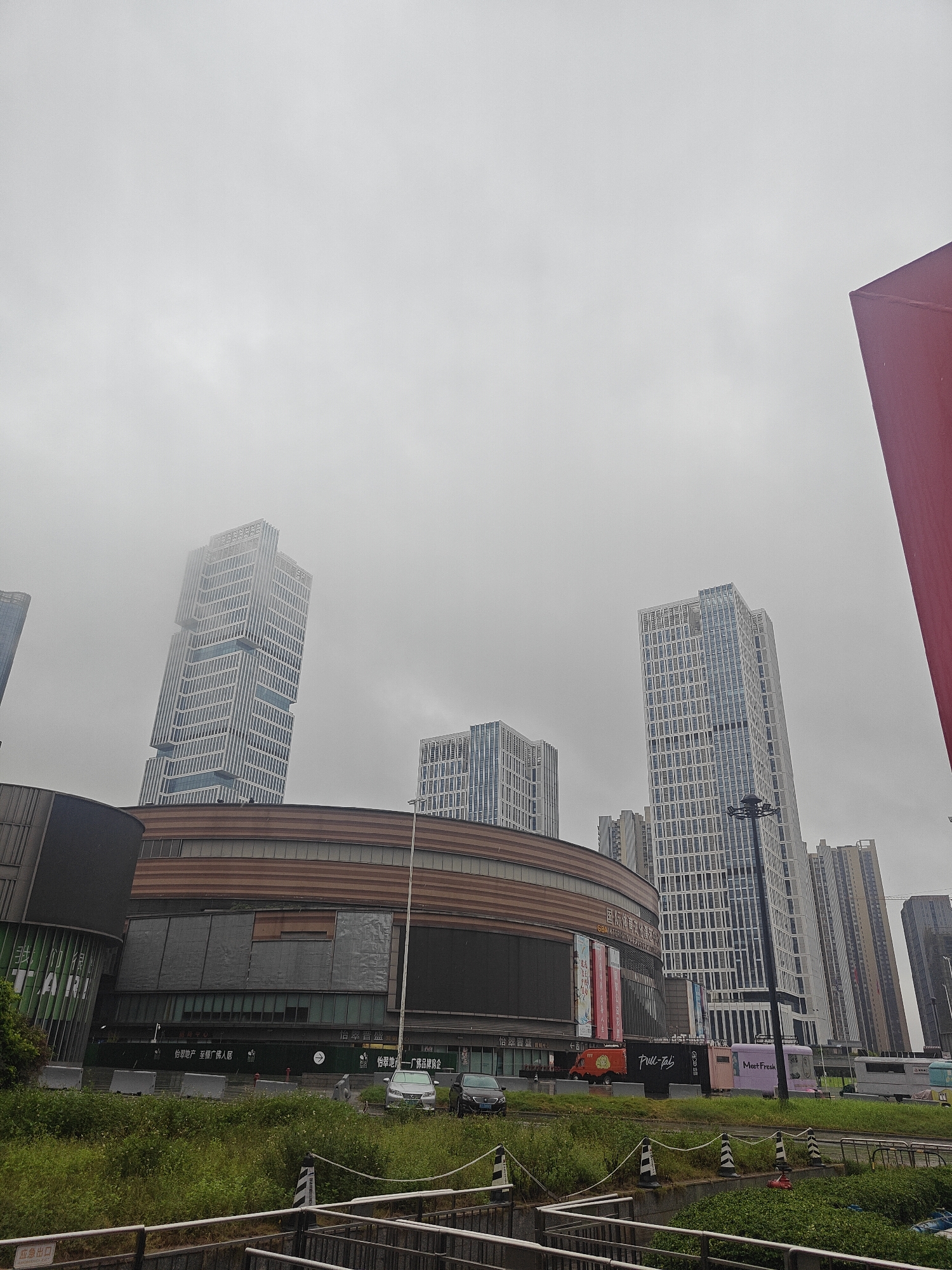 This media file shows a street view of a modern urban environment in Shunde District, China. The scene is dominated by several tall, contemporary buildings with glass facades, indicative of a commercial or business district. One distinctive skyscraper has a unique, stepped design, partially obscured by a light fog or haze, suggesting overcast weather. In the foreground, a large, curved building with a brown, textured exterior, possibly a shopping mall or entertainment complex, is visible. This building features large display screens and banners, one of which displays Chinese text and the word "TARI".
Below the large building, a row of food trucks and temporary structures are parked. One truck is labeled "Pull-Tab" and another "Meet Fresh", suggesting a food or beverage vendor area. Chinese text on banners and signage indicates commercial activity, with phrases like "怡翠地产—广佛品牌房企" (Yicuiprod-Guangfo brand real estate enterprise) and "怡翠晋盛" (Yicuijin sheng). A green barrier with more Chinese text is present in front of the buildings.
To the left of the main structure, a sign with "应急出口" (Emergency Exit) is visible. A patch of green grass and vegetation lines the foreground, with metal railings and a concrete structure suggesting a pedestrian walkway or a drainage system. The overall atmosphere is muted, likely due to the cloudy or foggy weather, which softens the sharp lines of the architecture. No people are prominently visible, giving the impression of a quiet moment in a bustling urban setting. The time of day appears to be daytime, given the ambient light.