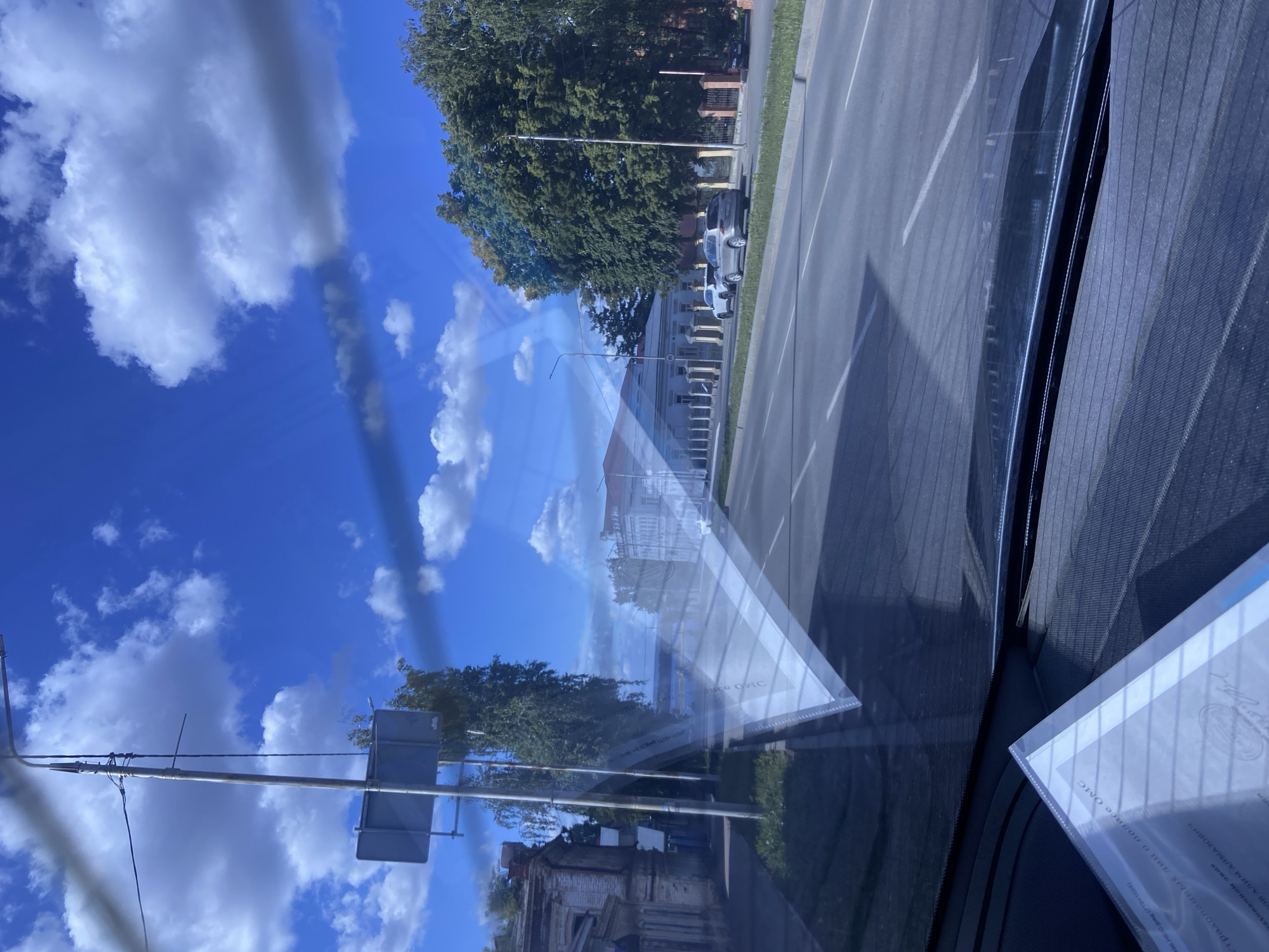 The image presents a sunlit street scene in Chistopol, Russia, captured from the interior of a vehicle, likely through a window or windshield. The perspective includes significant reflections, superimposing elements of the sky and documents onto the external view.
In the immediate foreground on the right, parts of the vehicle's interior are visible, including a textured, dark surface—possibly a sunshade or speaker cover—and two transparent plastic folders containing documents. These documents are partially readable through the reflections.
Beyond the reflections, the outdoor scene unfolds under a bright blue sky dotted with scattered white cumulus clouds. A paved road or street with white lane markings stretches across the midground, with parked cars visible along its side. A dense line of mature green trees borders the left side of the street, partially obscuring buildings behind them. Utility poles and street signs are also discernible.
The time of day is bright and sunny, indicative of fair weather. The documents within the folders display Russian text, including phrases such as "трахованных лиц о полисе Омс" (insured persons about the OMS policy) and "рахованном лице" (insured person), along with the name "ОВ АЛИМ АЛМАЗОВИЧ". These indicate medical insurance papers. The prominent reflections add a layered and somewhat abstract quality to the visual composition.