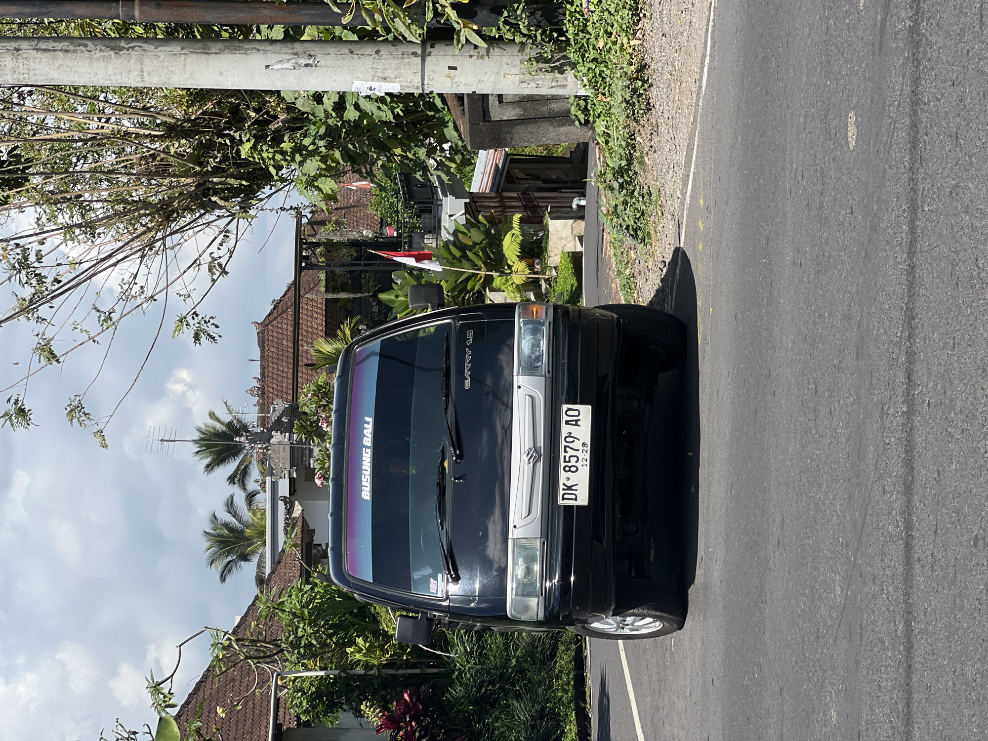 The image shows a black Suzuki Carry utility vehicle parked on the side of a road in Payangan, Indonesia. The vehicle has a license plate that reads "DK 8579 AO". A sticker on the windshield displays the text "BUSUNG BALI". In the background, there are traditional Balinese houses with tiled roofs, lush green vegetation, and palm trees. A Indonesian flag is visible on a pole. A utility pole stands to the right of the vehicle. The sky is partly cloudy.