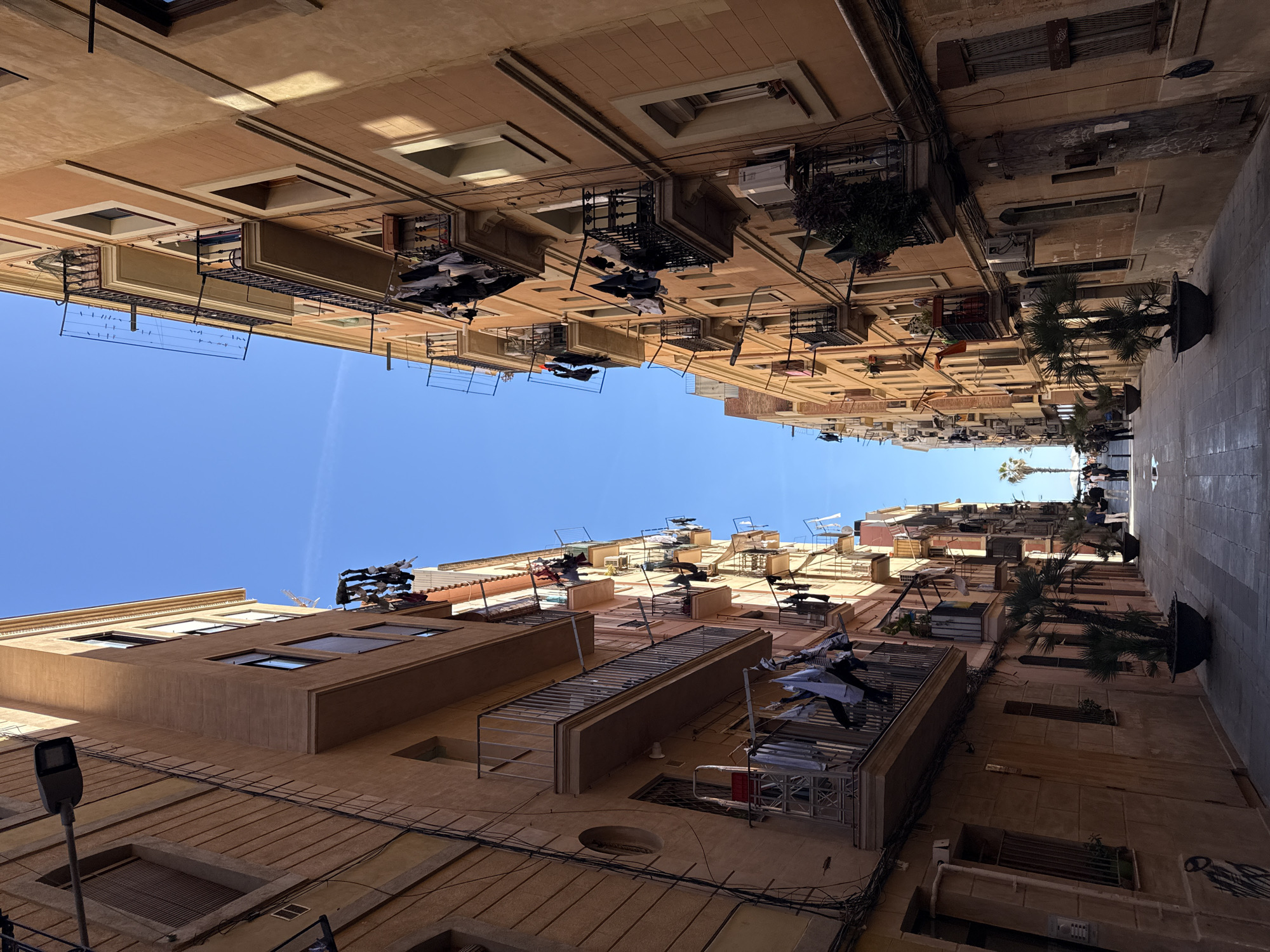 This is a street view looking up in Barcelona, Spain. The buildings are tall and closely spaced, creating a narrow alleyway effect. The architecture features balconies, some of which are used for drying laundry. There are a few trees in pots along the street. In the distance, figures of people are visible. A clear blue sky is visible at the top of the frame.