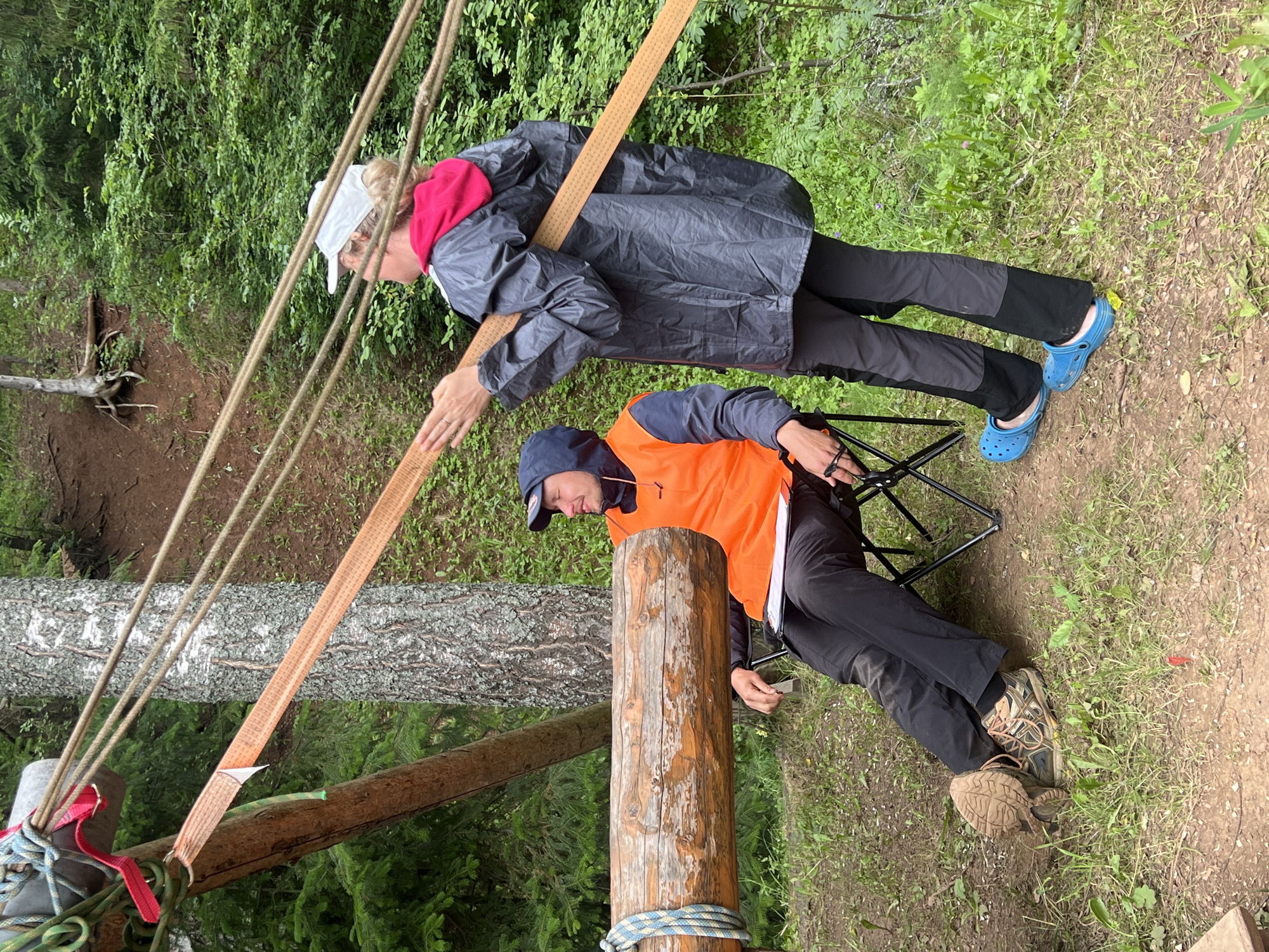 Two individuals are engaged in an outdoor activity in a wooded area. The person in the foreground is seated on a folding chair, wearing an orange safety vest, black pants, and hiking boots. Their head is tilted down, and they appear to be holding something small in their hand. The second person, standing behind and to the left of the first, is wearing a grey raincoat over a red hoodie, black pants, and blue Crocs. They are leaning forward, with one hand resting on a wooden beam, and seem to be looking down.
The scene is set in a forest with lush green foliage and trees. Several ropes and a wide, orange strap are visible, suggesting some kind of setup, possibly for an adventure activity like slacklining or a rope course. A large wooden log is positioned prominently in the foreground, with rope tied around it. Another fallen tree trunk is visible in the background. The ground is covered with dirt and grass. The overall impression is that of a daytime activity, possibly during overcast weather given the muted lighting. No visible text is present in the image. The location is identified as Sergino, Russia.