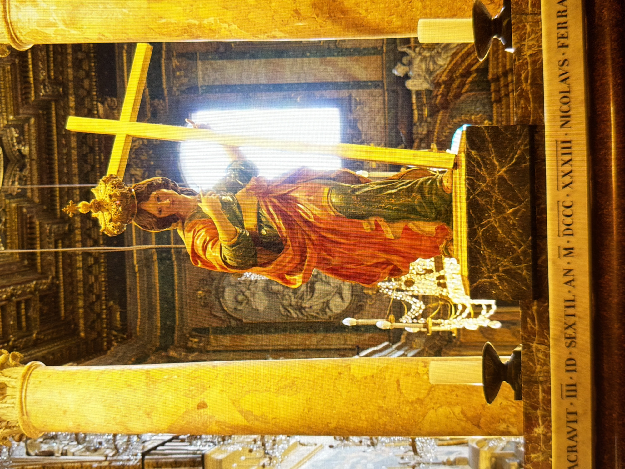 The image shows a statue of a female figure wearing a crown and holding a large golden cross. The statue is positioned in what appears to be a church or religious building, with ornate columns and architectural details visible in the background. There are also candles and what looks like an altar or pedestal in the foreground. The lighting is warm and golden, possibly from artificial lights or sunlight filtering through a window. The statue's clothing is draped in rich red and green fabric, and the crown is elaborate and gilded. A band with inscriptions in Latin is visible at the bottom of the image. The scene is focused on the religious iconography and the grandeur of the setting. There is no indication of the location being in Kyiv, Ukraine.