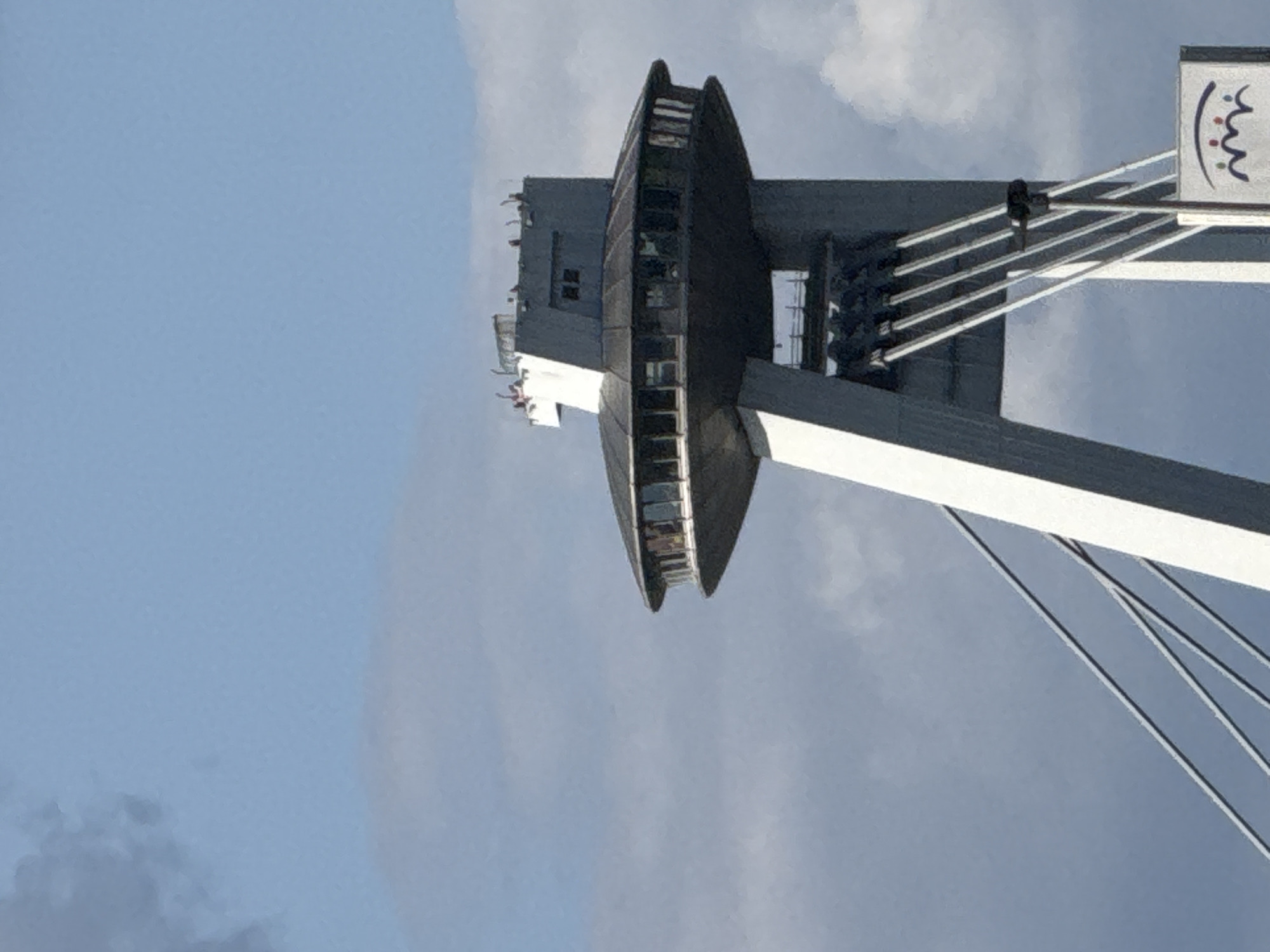 The image shows the UFO Observation Deck, a prominent landmark in Bratislava, Slovakia, situated atop the SNP Bridge. The structure is a modern, disc-shaped building with large windows, elevated on a concrete pylon. Several people are visible on the observation deck, appearing to be visitors enjoying the view. The sky is partly cloudy, with a mix of blue sky and white clouds. Cables extend from the pylon, supporting the observation deck, and a sign with an abstract logo is visible at the base of the structure.