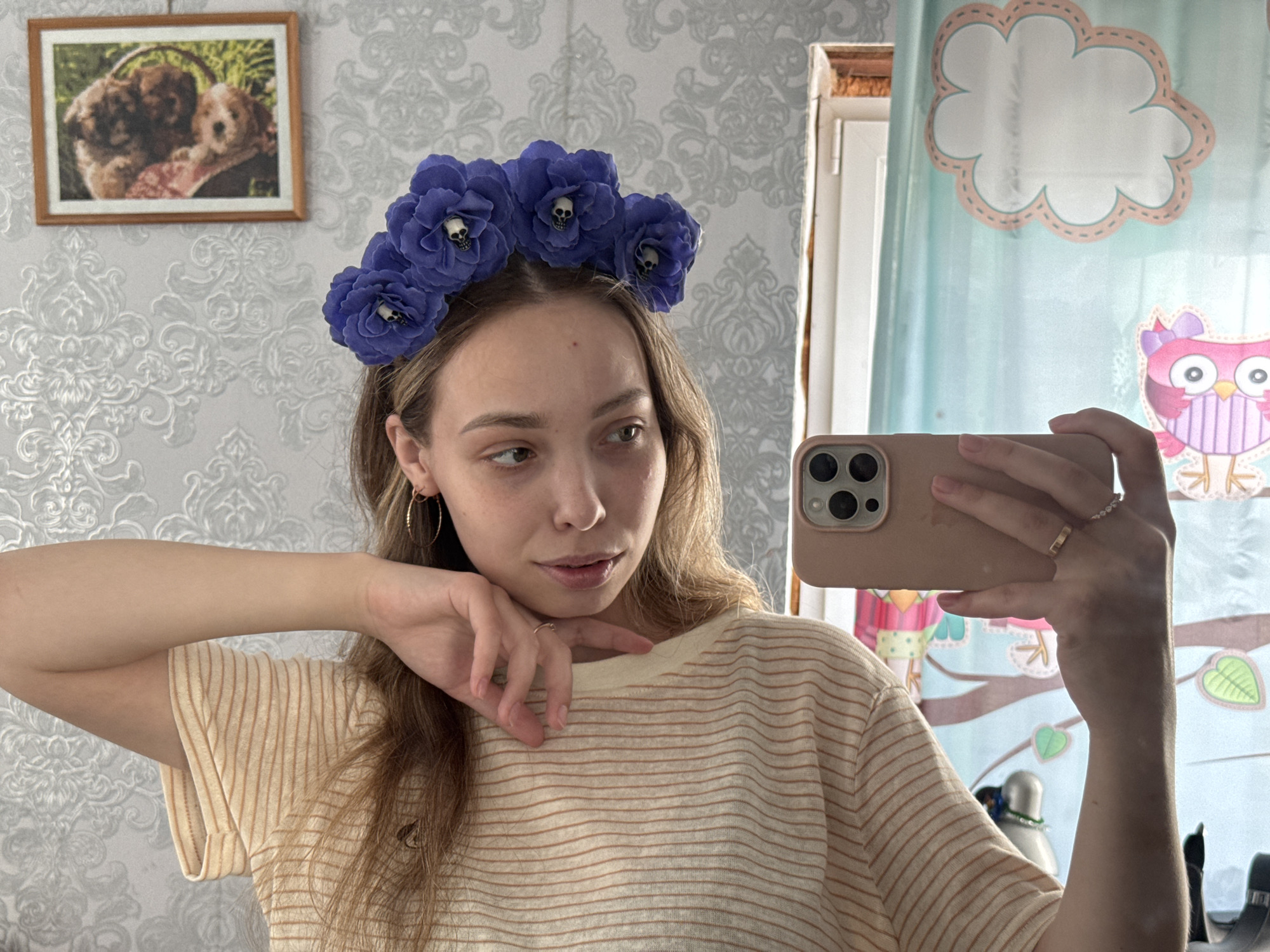 A woman is taking a selfie in a room. She is wearing a light yellow and brown striped shirt, gold hoop earrings, and a blue floral headband with small skull accents. She is holding a light pink smartphone in her right hand, capturing her reflection in a mirror. Her left hand is resting under her chin.
The room has wallpaper with a light grey and white floral pattern. A framed picture of two puppies hangs on the wall. To the right of the woman, a curtain with a cartoon-like owl and cloud design is visible. Other objects are partially visible in the mirror, including a white cylindrical object with what appears to be a lamp or light fixture. The specific location in Кемуль, Russia is not discernible from the image.