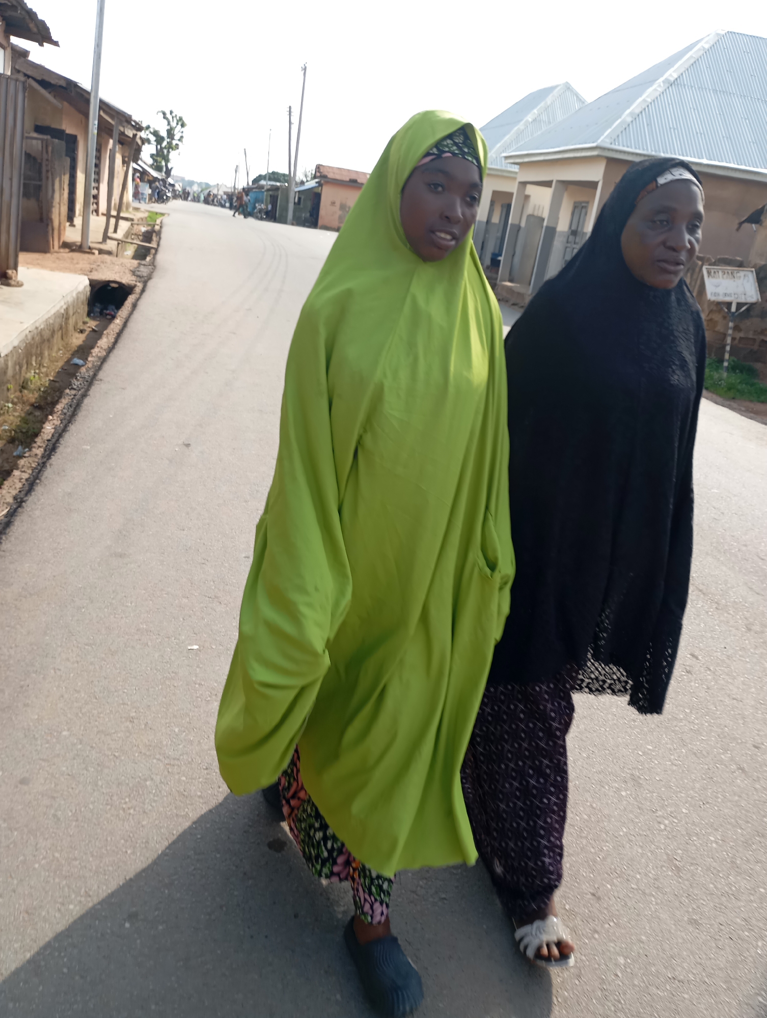 Two women, one wearing a bright green hijab and the other a black hijab, walk side-by-side on a paved road in Shendam, Nigeria. The woman in green has a patterned headscarf visible under her hijab and wears a dark, patterned skirt and dark shoes. The woman in black wears a black patterned skirt and white sandals. They are walking towards the viewer, with the road extending into the background. The surroundings suggest a residential or commercial area, with buildings and a street lined with what appear to be shops or houses. A drainage ditch runs alongside the road on the left. The sky is overcast, and the lighting suggests it is daytime.