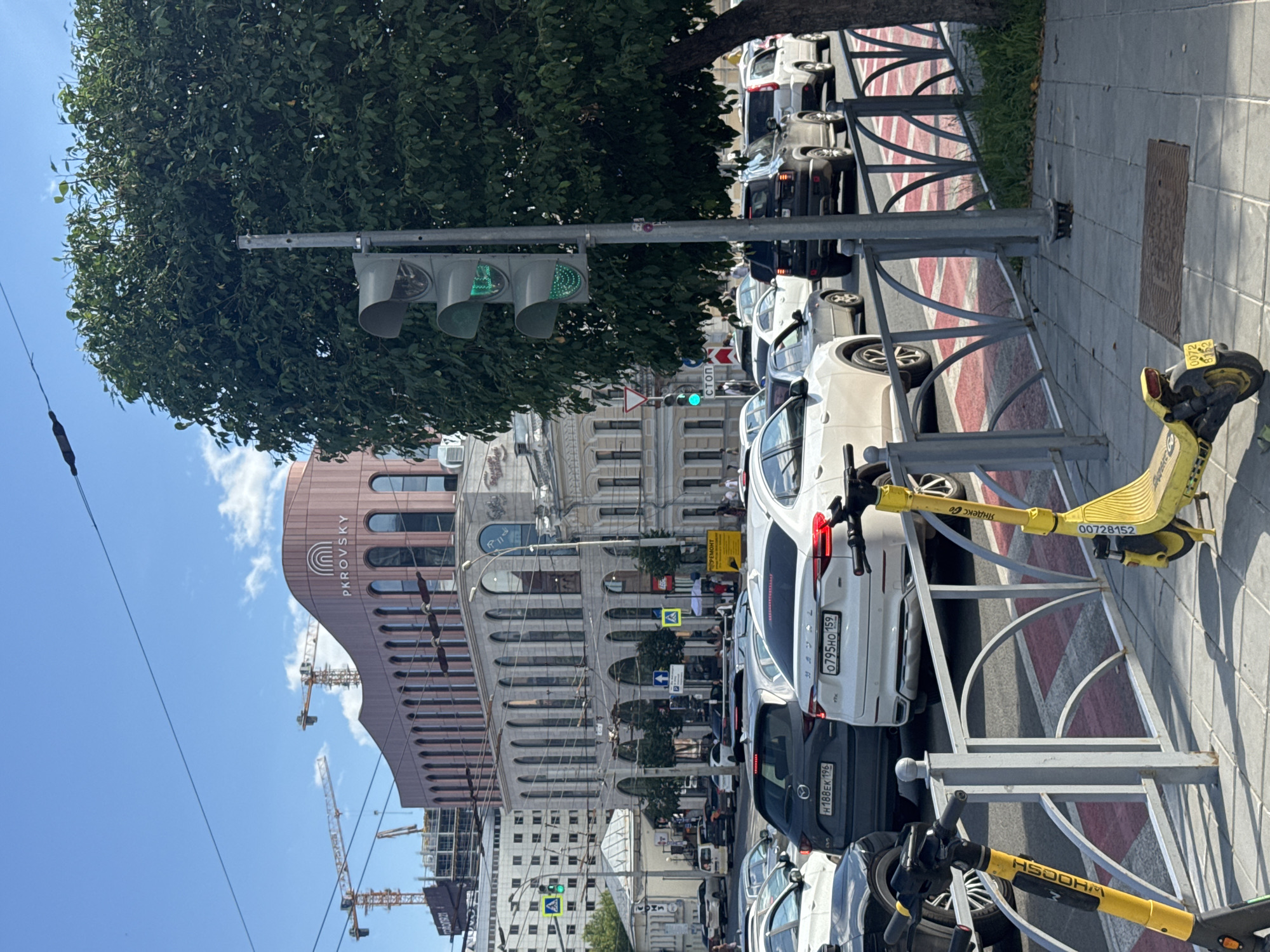 The image captures a street scene in Yekaterinburg, Russia, focusing on traffic and urban architecture. A traffic light with a green signal is prominently displayed on the right, amidst the dense foliage of a large tree. In the foreground, two yellow electric scooters are parked near a metal fence. The middle ground is dominated by a moderate traffic jam on a city street, with various cars including a white Haval, a dark grey Mazda, and other vehicles visible. Behind the traffic, an architecturally distinctive building with arched windows and the signage "PKROVSKY" is visible. Construction cranes are present in the background, indicating ongoing development. The sky is clear with some scattered clouds. The overall environment suggests a sunny day in a busy urban area.