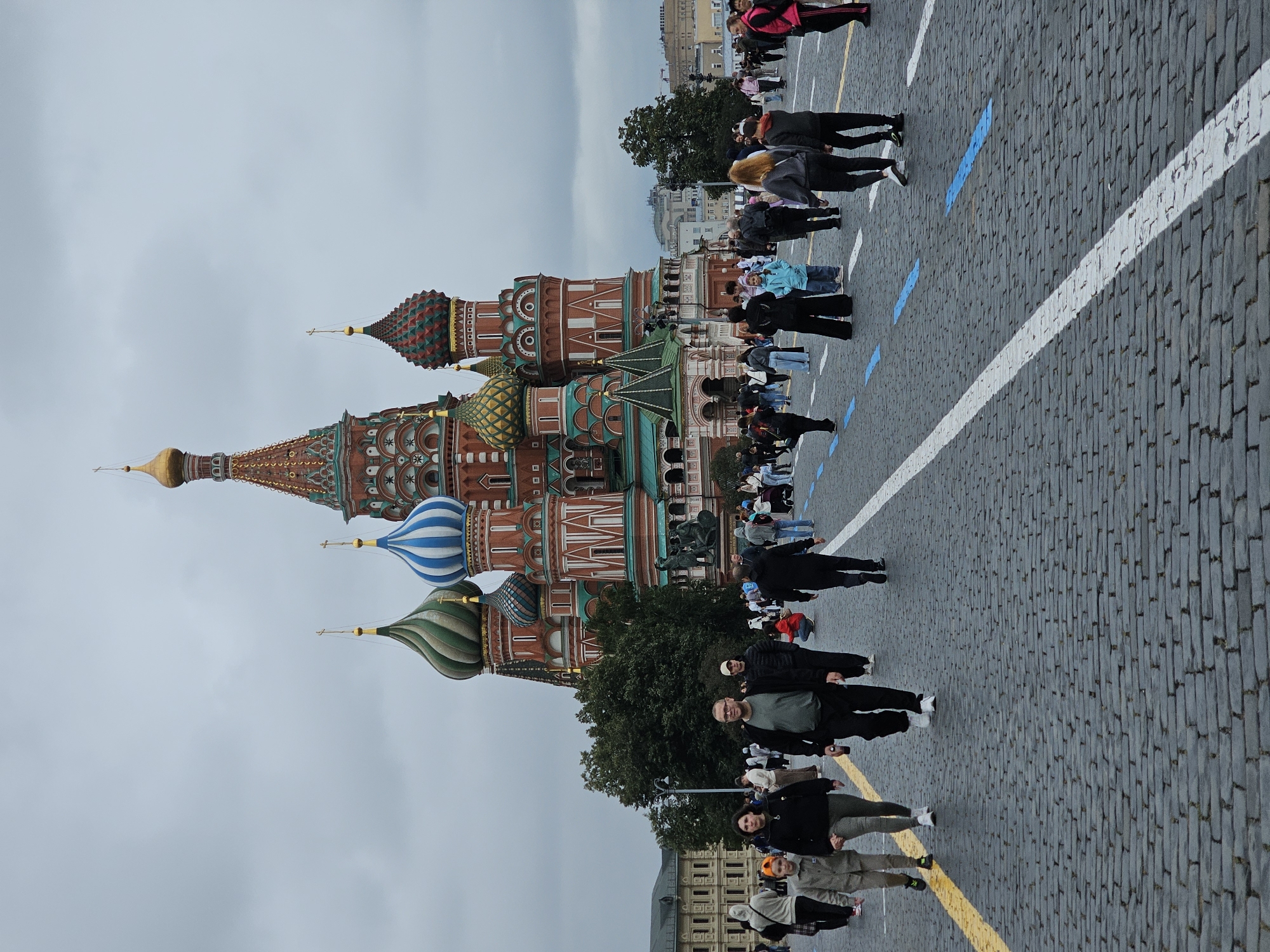 A high-angle, full shot captures the vibrant St. Basil's Cathedral in Moscow, Russia, under an overcast sky. The iconic onion domes, painted in vivid red, green, and blue, dominate the background. In the foreground, a cobblestone plaza is filled with pedestrians walking in various directions. The paving stones are wet, suggesting recent rain. A yellow line runs along the left side of the plaza, and white lines with blue rectangular markings create a delineated path. People are dressed in casual clothing, some in jackets and hoodies, indicating cooler weather. A group of three young people are walking towards the camera on the left side, with a man in the center looking directly at the camera. Other individuals are scattered throughout the plaza, engaged in walking and conversing. The overall scene depicts a bustling public space with a prominent architectural landmark.