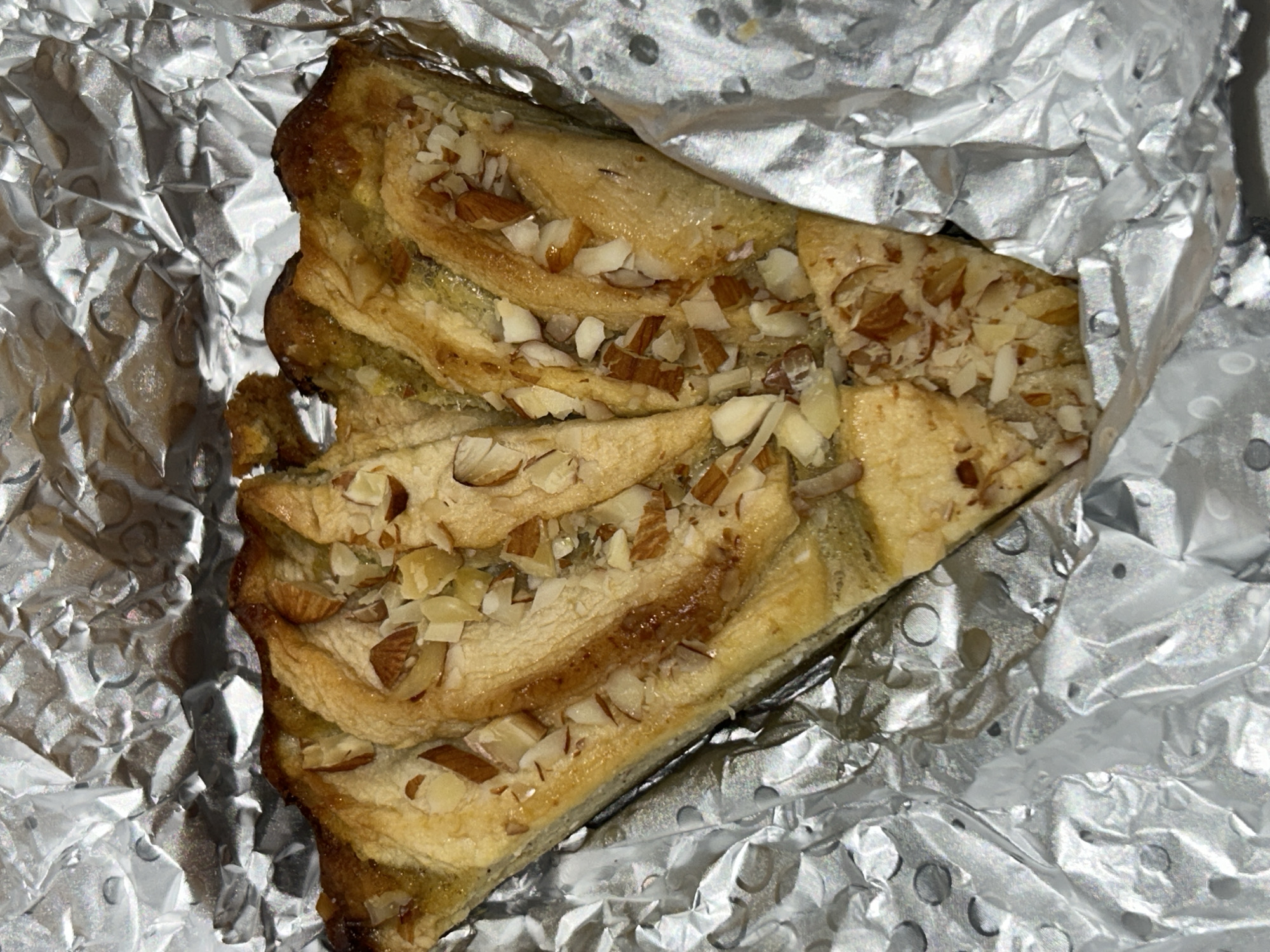 A partially eaten slice of baklava-like pastry sits on silver foil. The pastry is triangular and flaky, with visible layers, nuts, and a glaze. The silver foil is crumpled and reflects light unevenly.