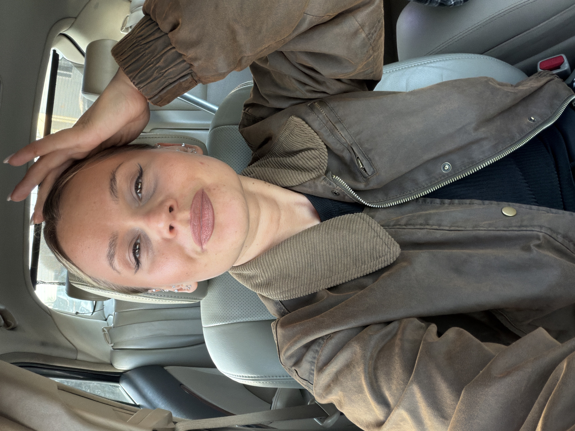 A woman is in the driver's seat of a car, wearing a brown jacket and a black shirt. She has fair skin, light brown hair, and wears multiple earrings in her left ear. She has one hand raised to her forehead. The car's interior is light gray leather. The location appears to be inside the vehicle and the context provided states that the location is in the city of Sarapul, Russia.