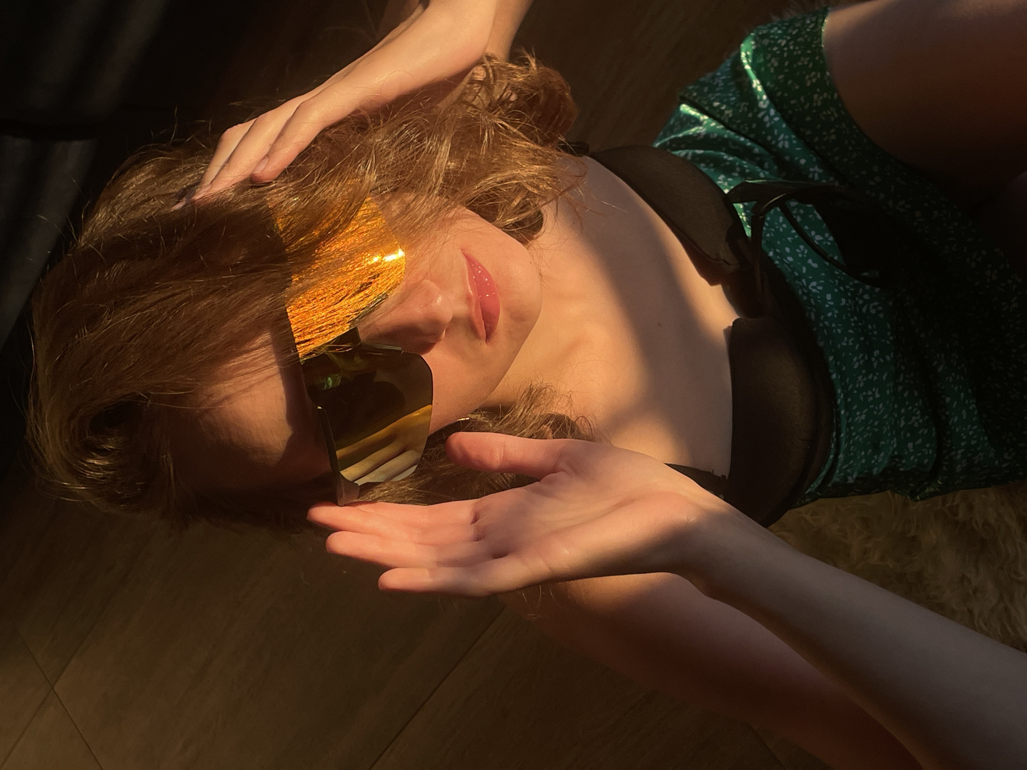 A person lies on a floor covered with wood planks. She wears a green floral dress with a black top underneath. Her hair is blonde and slightly tousled. She's wearing sunglasses with gold-tinted lenses and holding her hand out. The scene is lit by sunlight. The location is not discernible from the image.