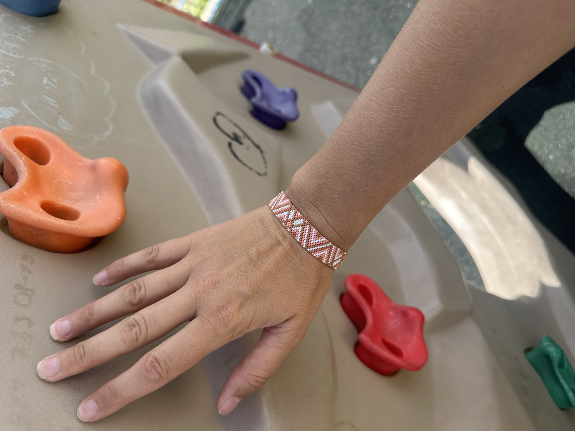 A person's left arm and hand are positioned on a light-colored climbing wall with red, orange, and purple climbing holds. The person is wearing a beaded bracelet with a geometric pattern of white, pink, and peachy-orange beads. The hand is resting on the wall, with fingers slightly curled. The background appears to be outdoors, with a hint of greenery and pavement visible. There is no identifiable information to place this image in Petropavlovsk-Kamchatsky, Russia.