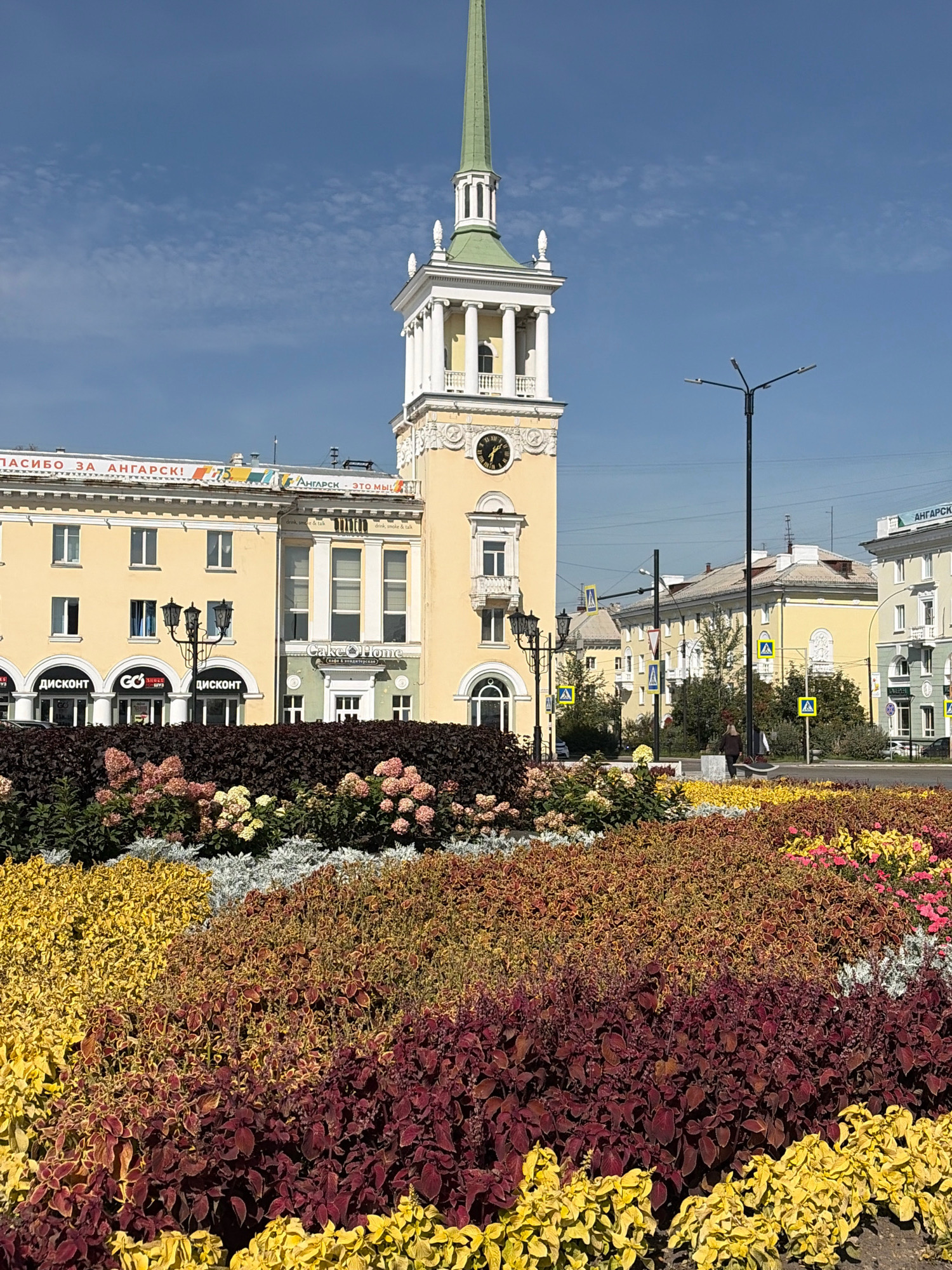This image captures a street scene in Angarsk, Russia, dominated by a prominent yellow building with a distinctive green-spired tower. The building features a clock on its facade and several arched windows on the ground floor, with smaller windows above. Signs on the building include "ДИСКОНТ" (discount) and "Cake & Home". In the foreground, a large, colorful flower bed displays a variety of plants in shades of yellow, burgundy, and pink, with some white and green foliage. A street lamp stands near the building, and a pedestrian crossing sign is visible in the background. The sky is clear blue with a few wispy clouds. A single person is seated on a bench in the middle ground.