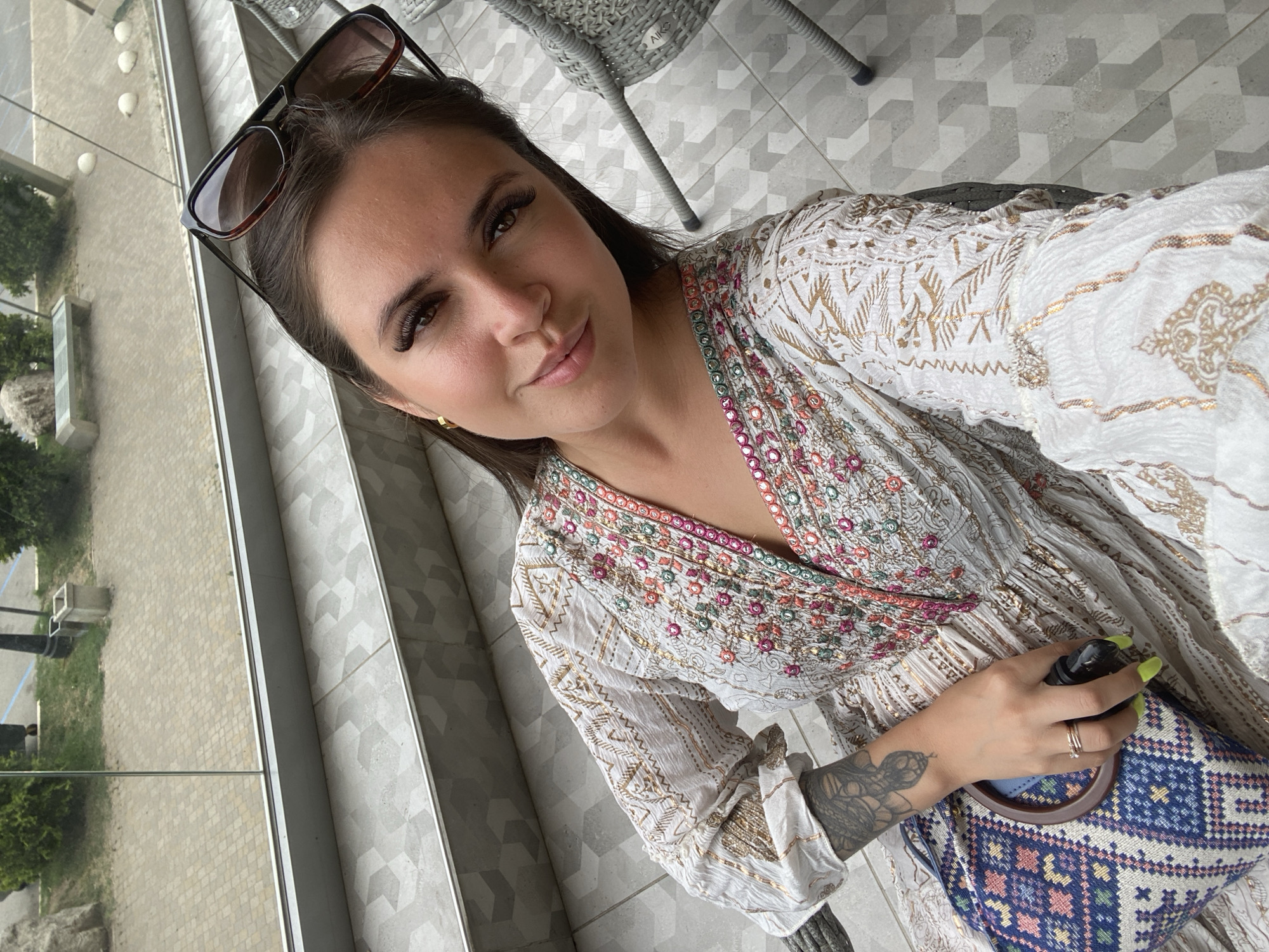 A woman is sitting at a table outdoors with a clear glass barrier in the foreground. She is wearing a white dress with intricate colorful embroidery and gold accents. She has sunglasses perched on top of her head and is holding a black object in her right hand. Her left arm is resting on a bag with a colorful geometric pattern. The background shows a courtyard with patterned paving stones and greenery. The location is the outdoor seating area of a restaurant or cafe in Derbent, Russia, offering a view of the surrounding urban landscape and park.