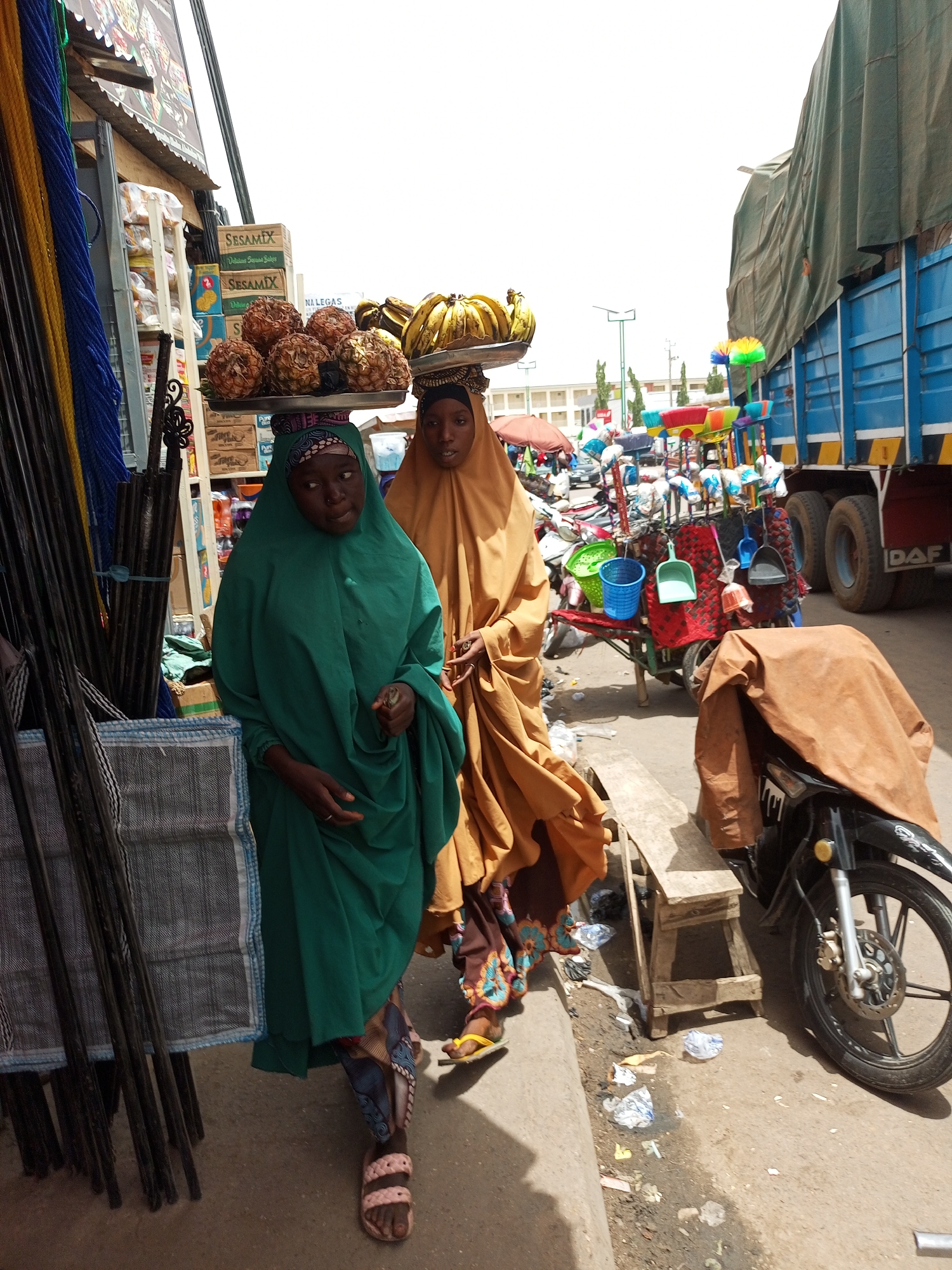 Two women in Arawa, Nigeria, are walking along a street with food trays balanced on their heads. The woman on the left is wearing a green robe and carrying pineapples, while the woman on the right is wearing a brown robe and carrying bananas. They are walking past a shop with goods displayed and a truck parked on the side of the road. A motorcycle is parked in front of the truck and a wheelbarrow carrying brushes and buckets is beside it. There is trash on the ground.